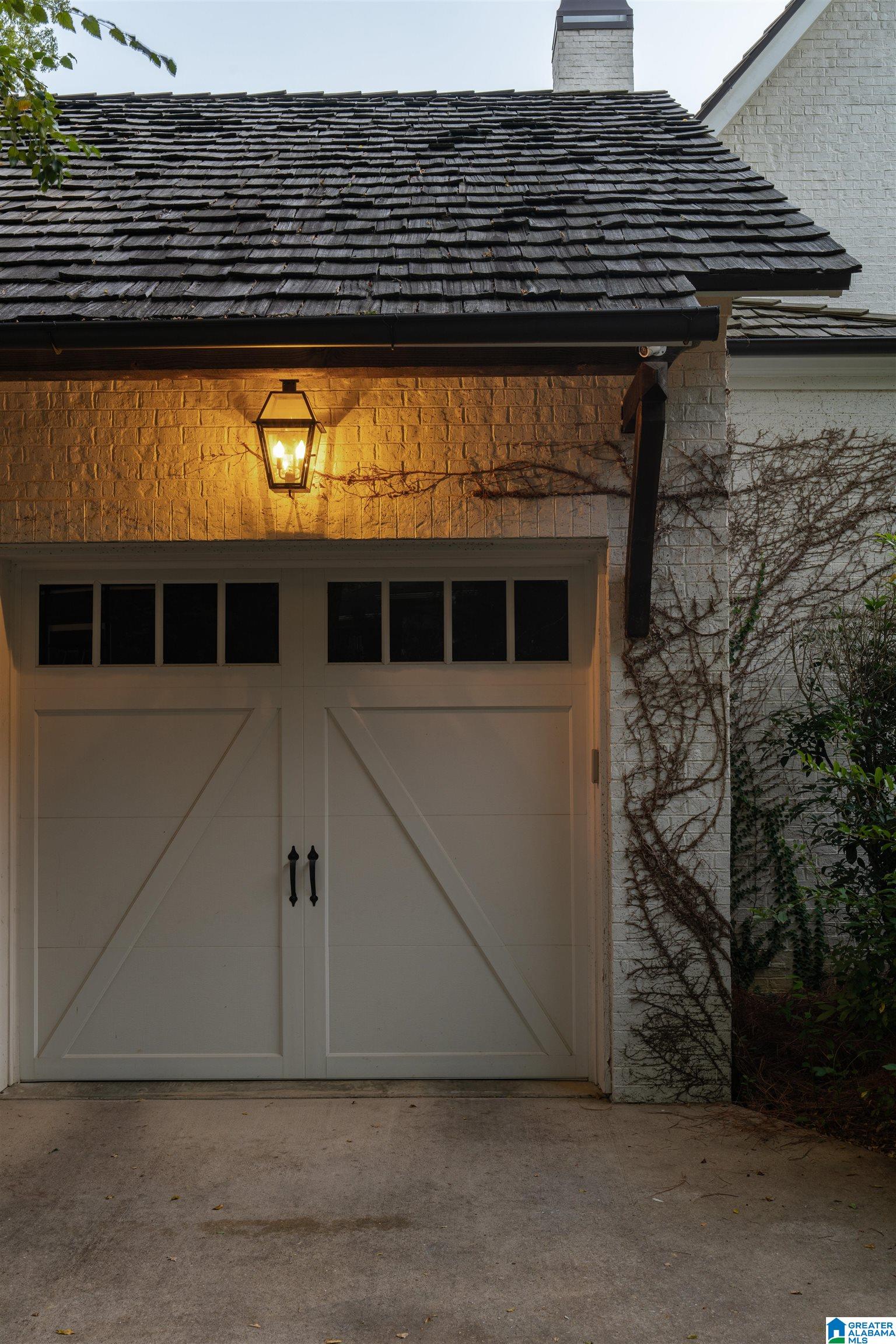 White brick garage with cream double doors, dark shingle roof, glowing lantern, and climbing vines.