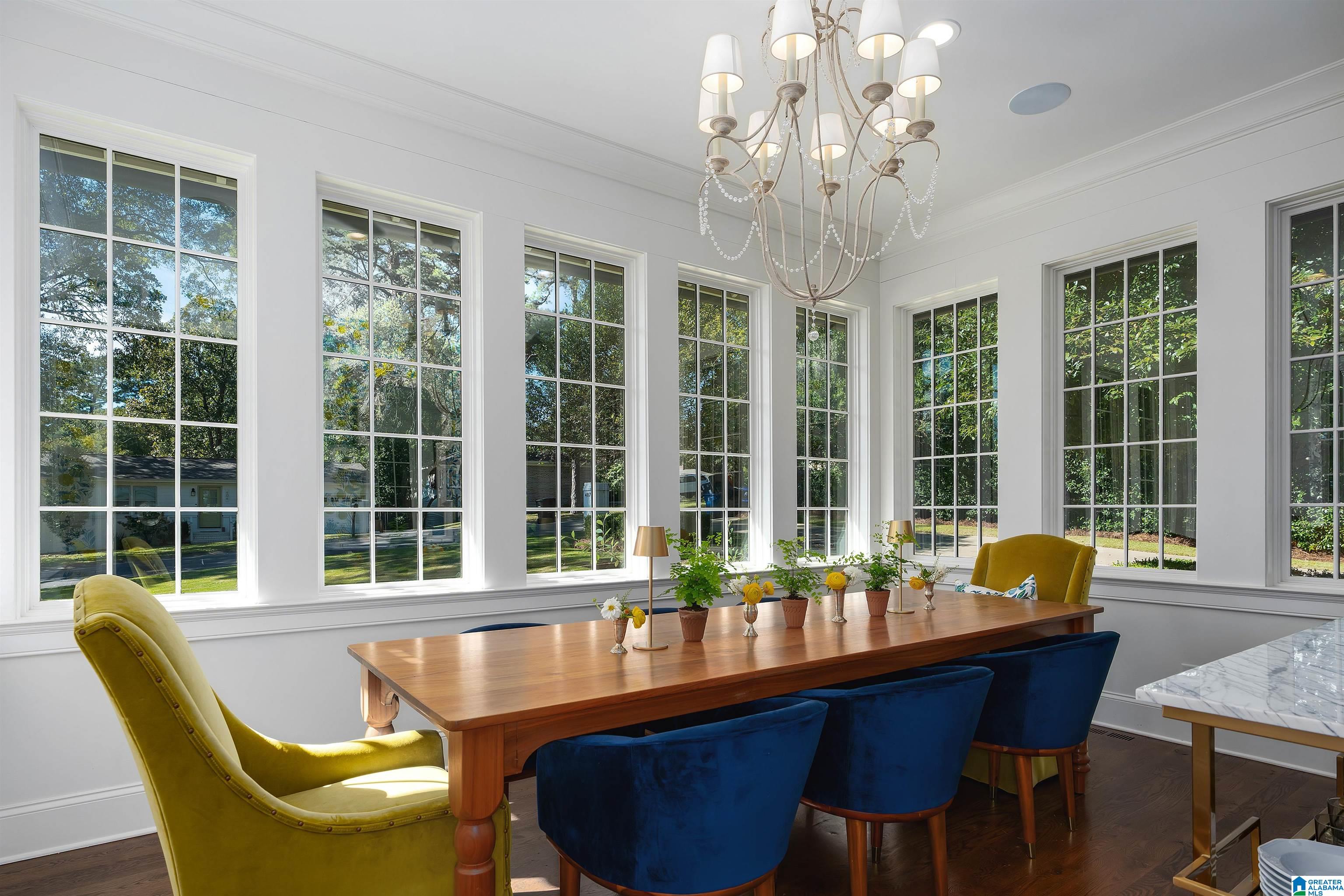 Dining room with a wood table, blue and yellow chairs, chandelier, and tall windows overlooking trees.