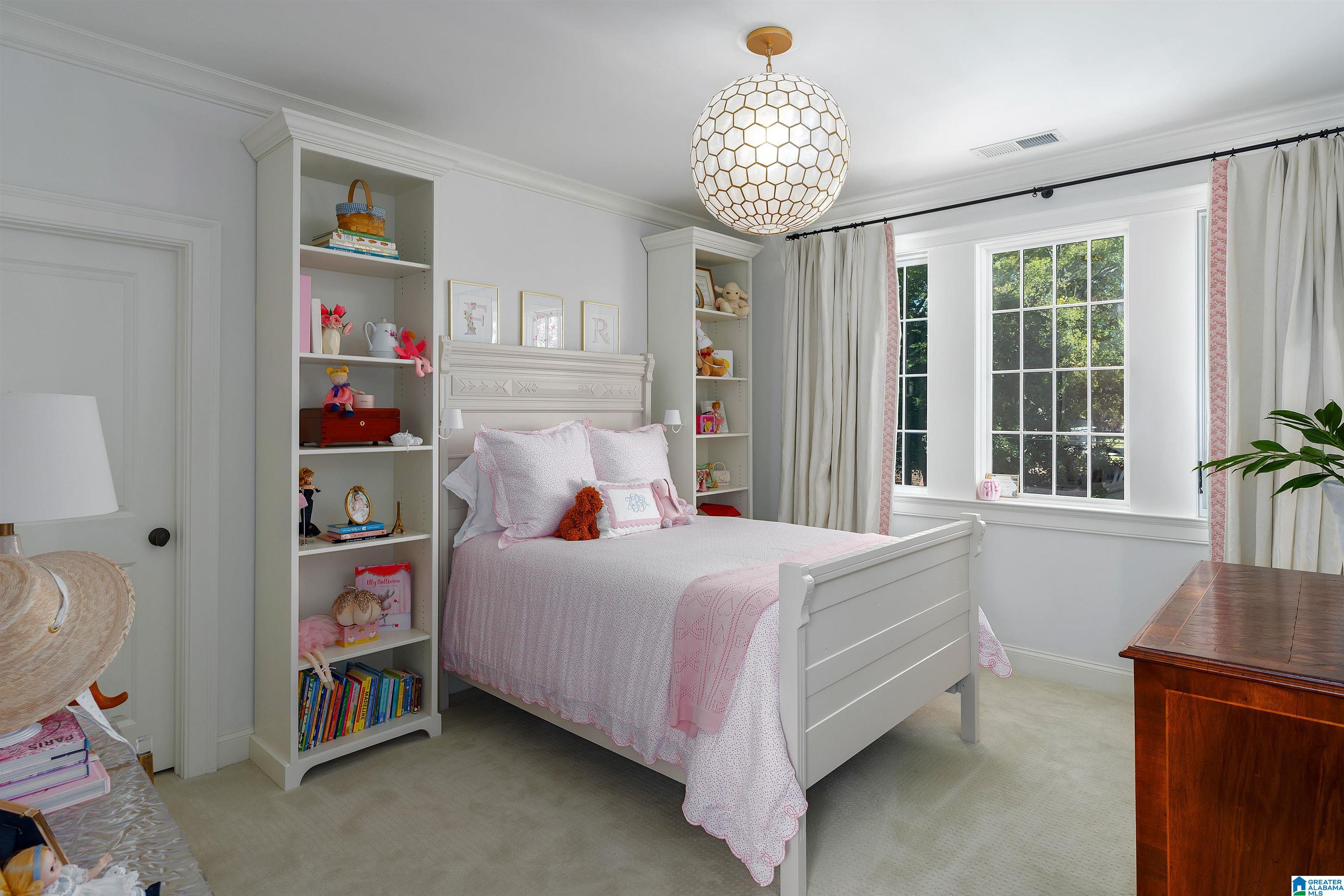 Bedroom with a white bed in pink bedding, toy-filled shelves, and a large sunlit window.