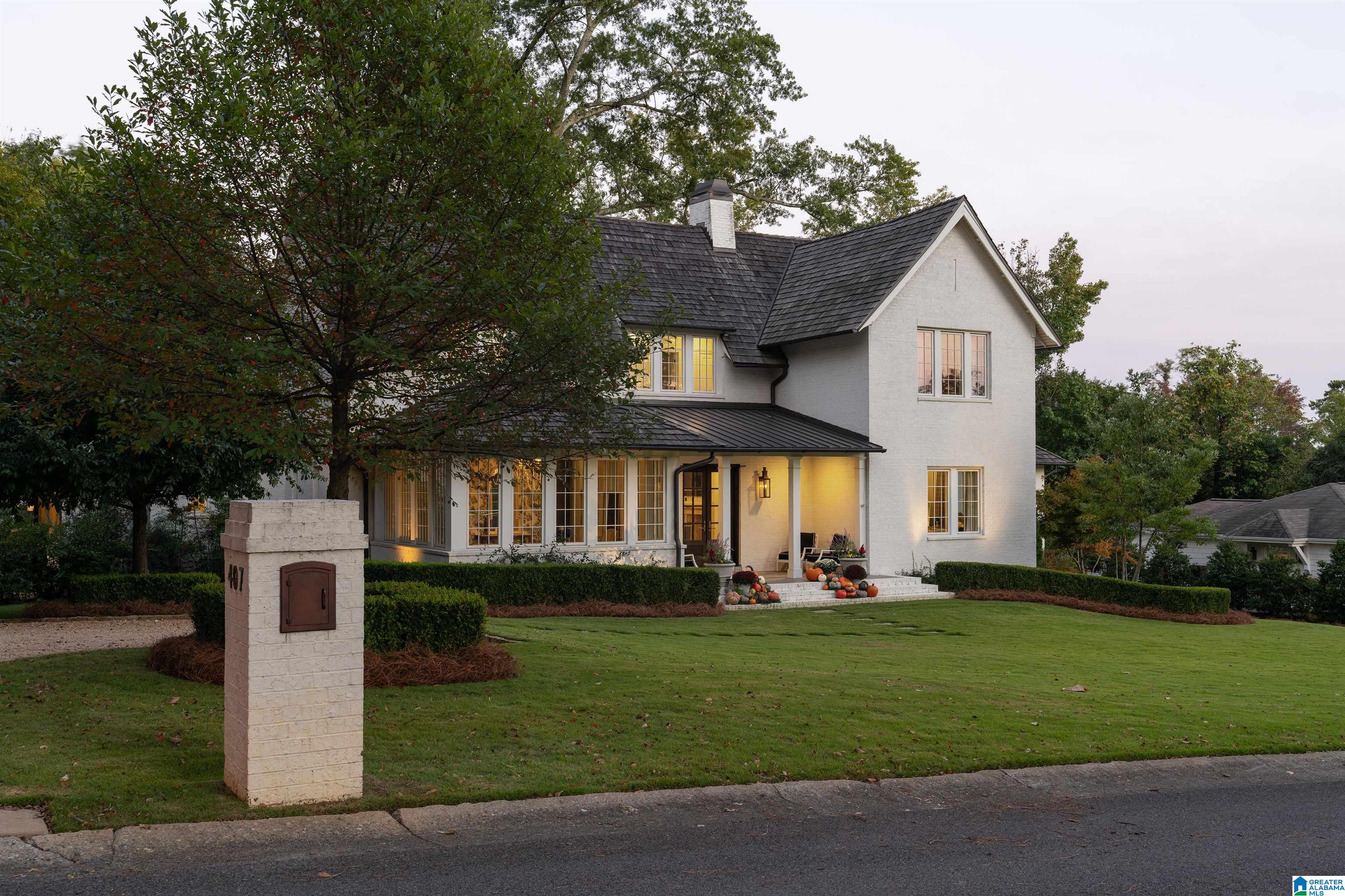 White two-story house with dark roofs, glowing windows, and pumpkins decorating the front steps.