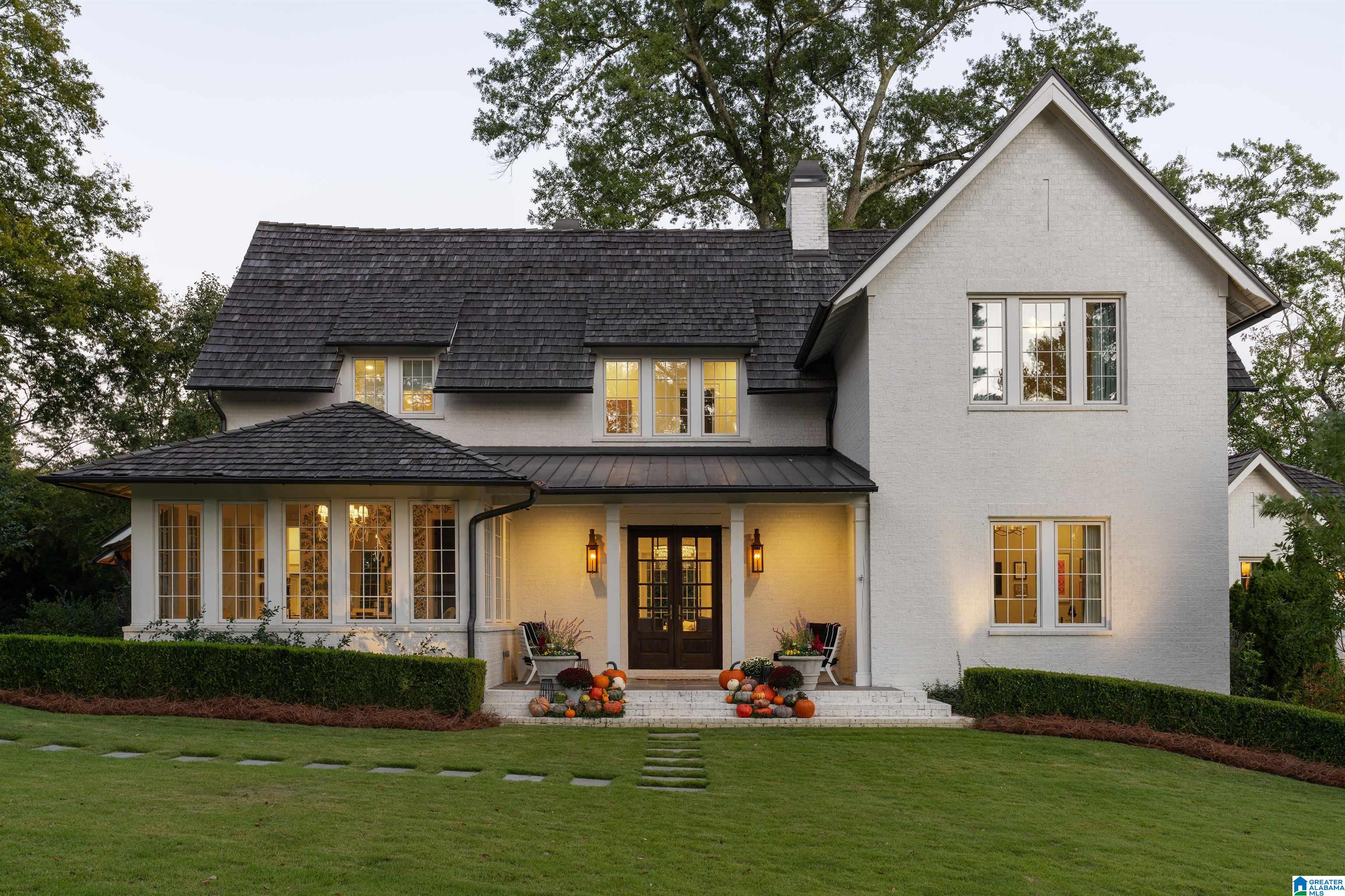 White two-story brick home with a dark roof, warmly lit windows, and pumpkins by the front entrance.