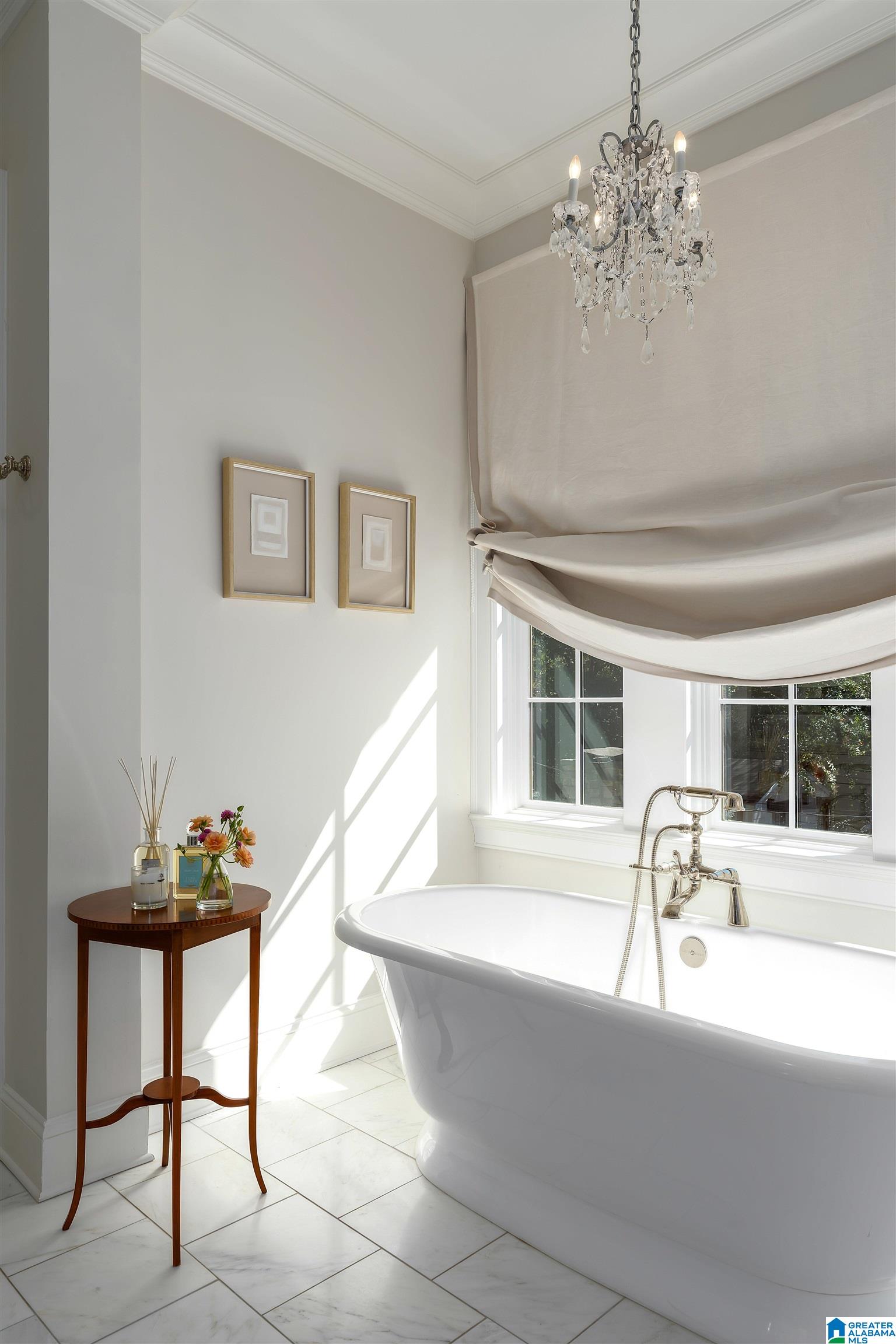 Bright bathroom with freestanding tub, crystal chandelier, and window with a Roman shade.