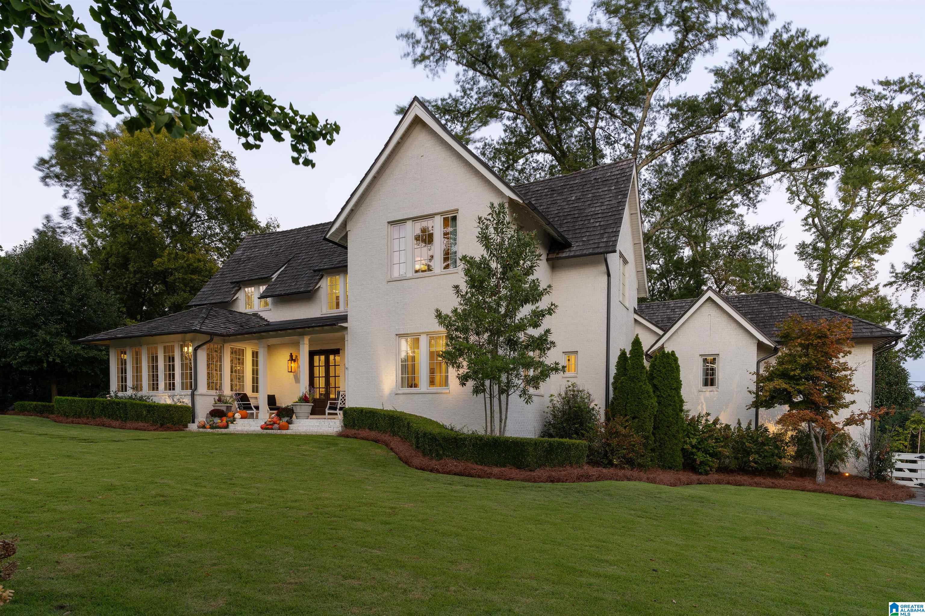 White-brick house with steep gabled roofs, a covered porch, glowing windows, and a broad lawn at dusk.