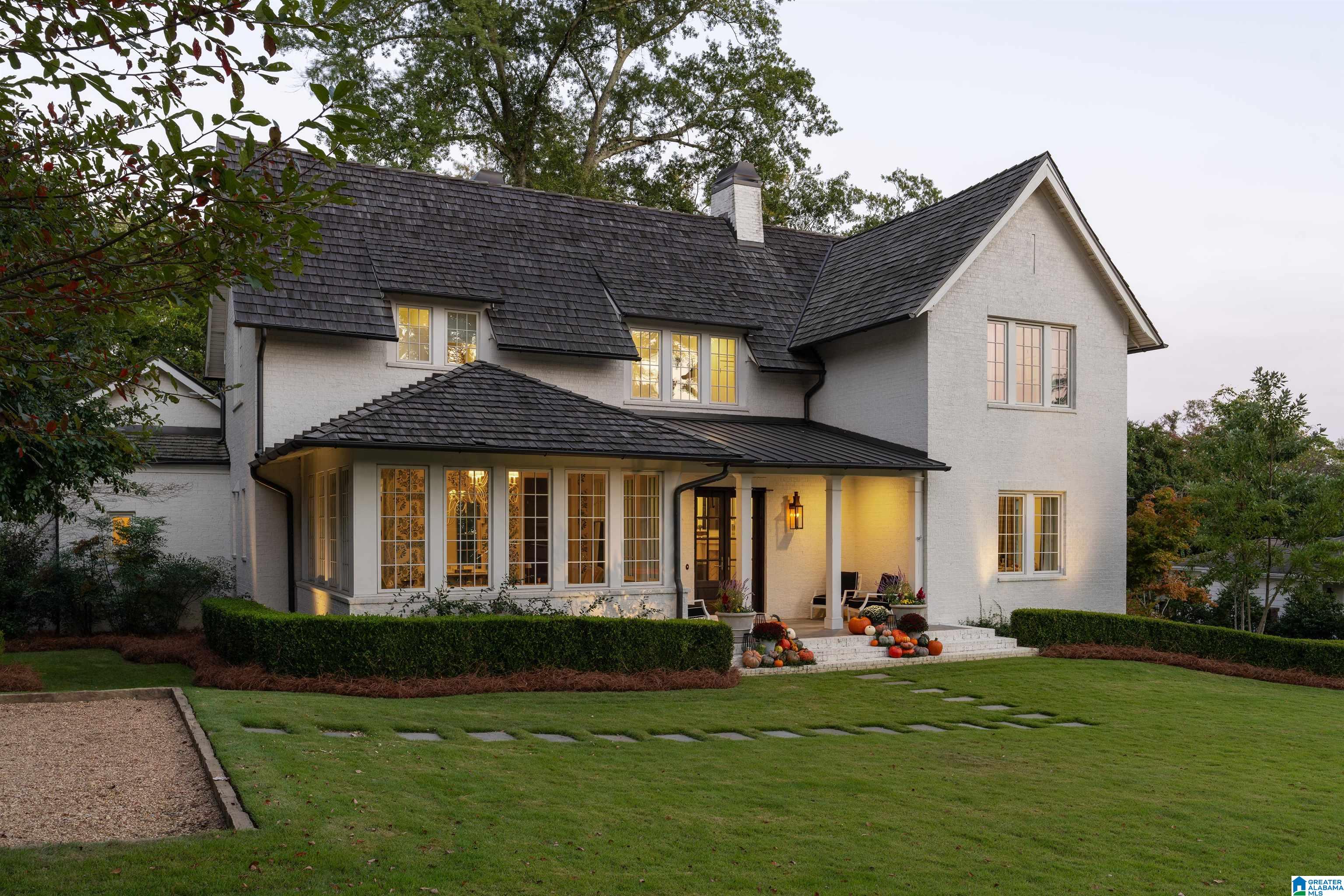 White brick home with dark tiled roofs, glowing windows, and pumpkins by the front porch.