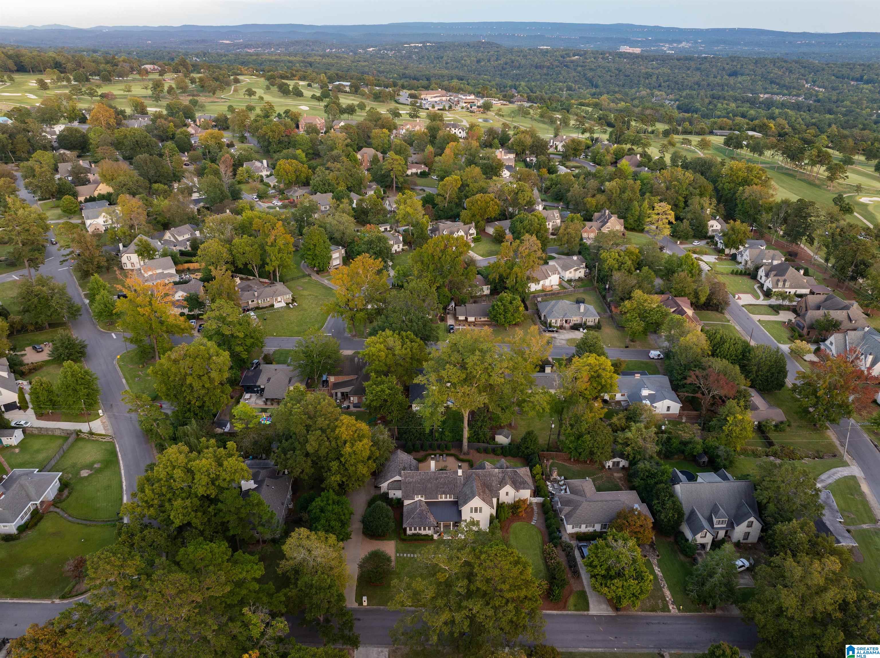 Aerial view of a leafy neighborhood beside a golf course, with wooded hills on the horizon.