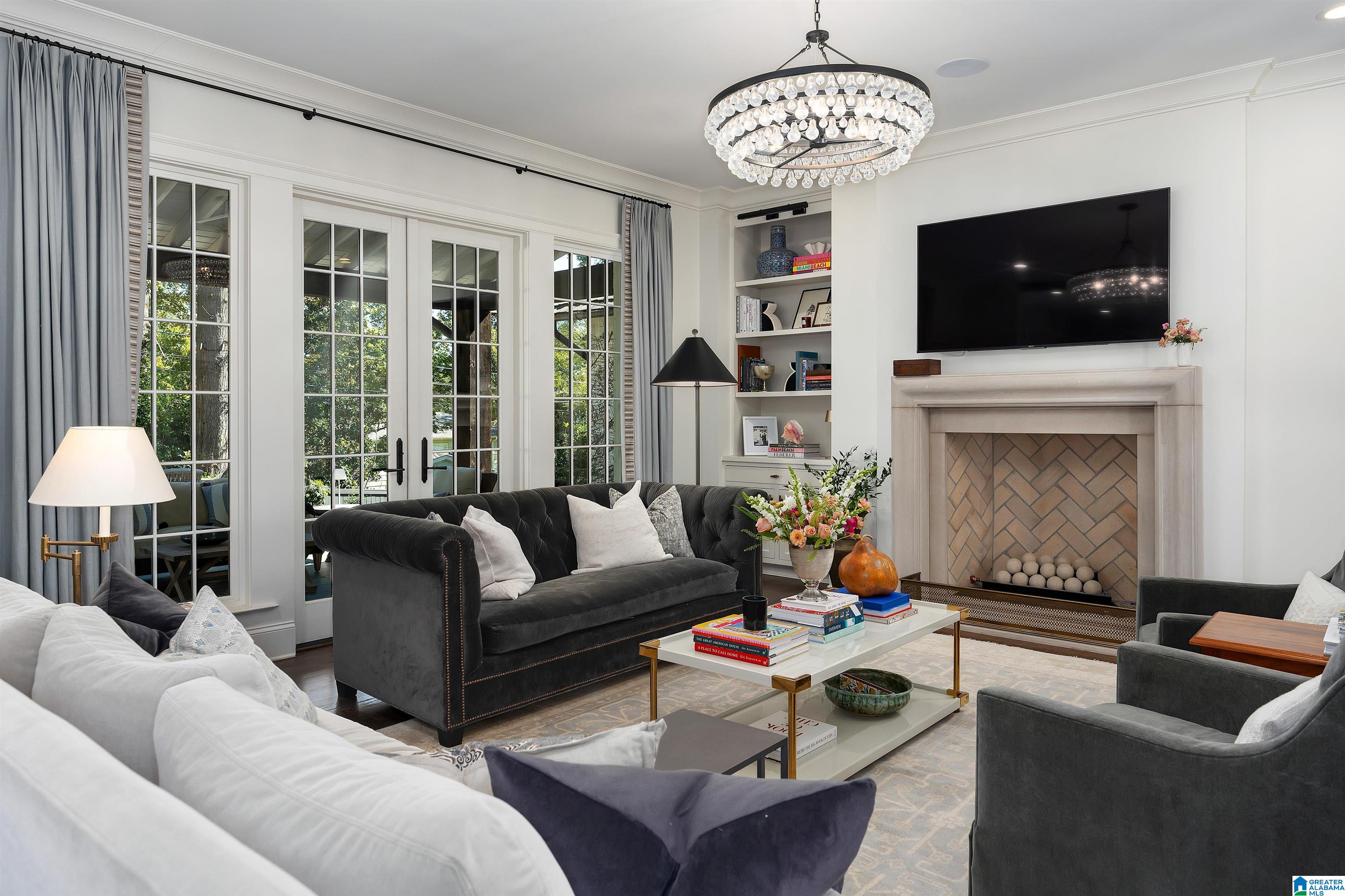 Living room with a stone fireplace, dark sofas, French doors, and a crystal chandelier.