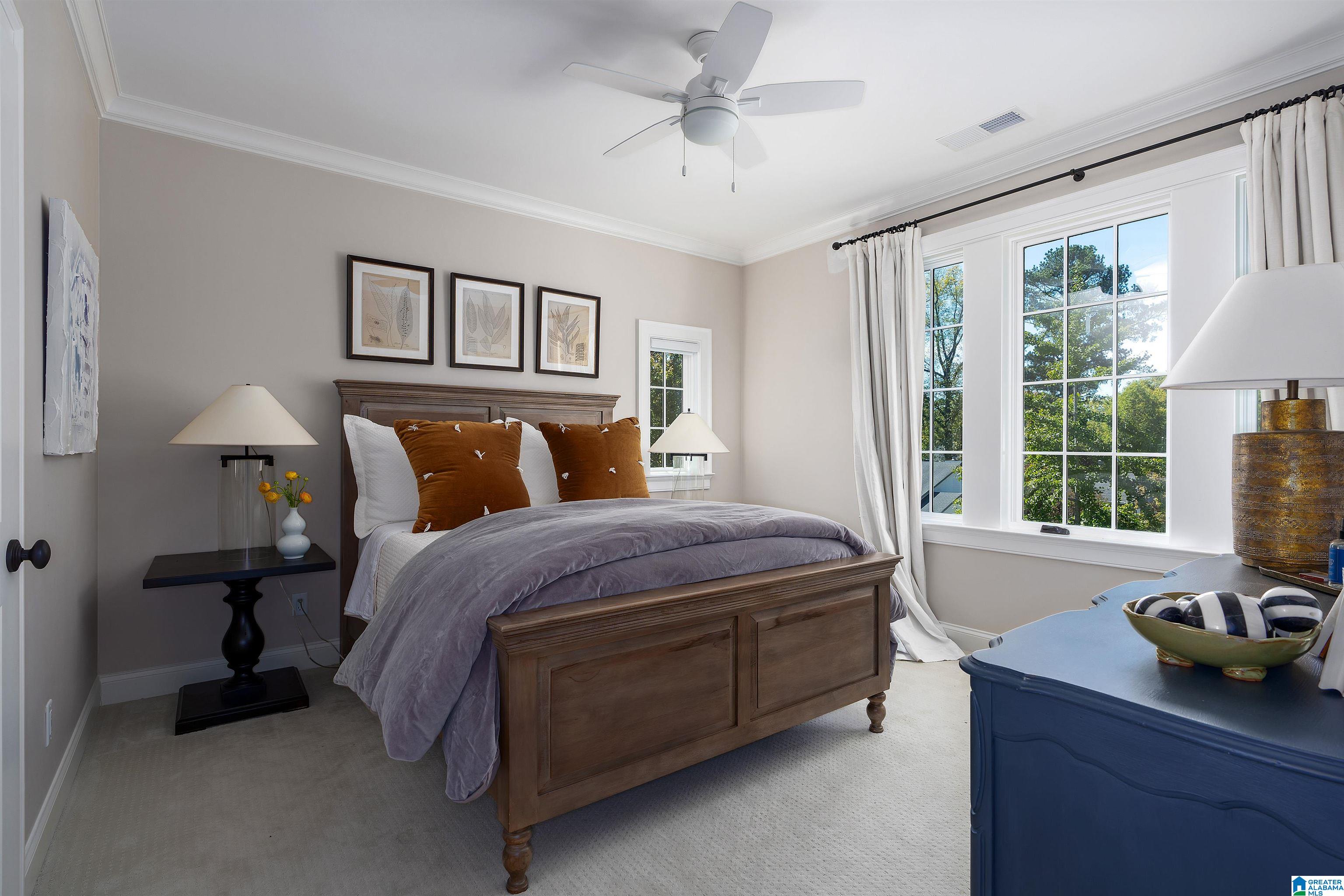 Bedroom with a dark wood bed, rust-colored pillows, botanical prints, and tall windows overlooking trees.