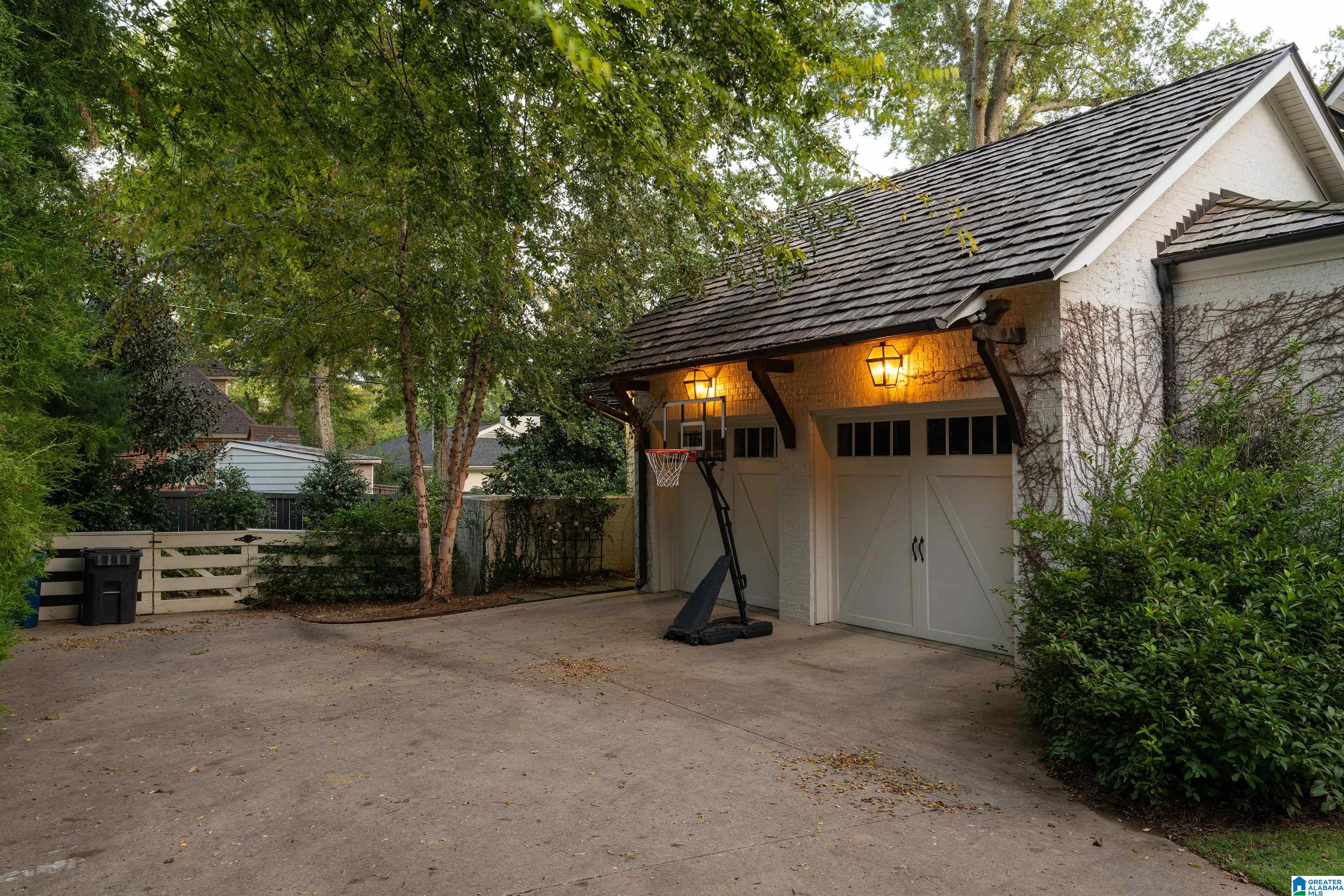 White brick garage with a steep shingle roof, lit lanterns, and a basketball hoop on the driveway.