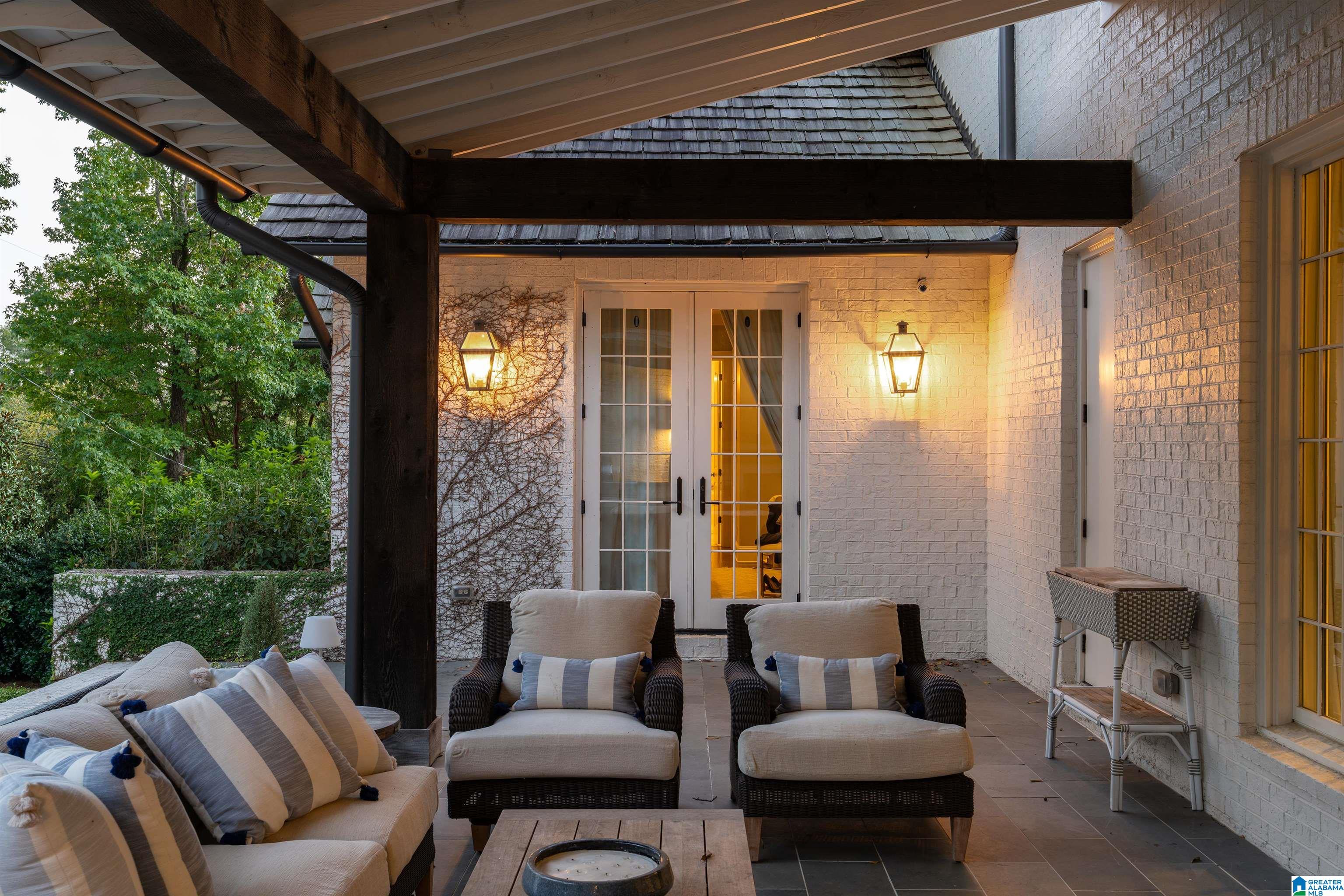 Covered patio with cushioned wicker seating, white brick walls, French doors, and glowing lanterns.