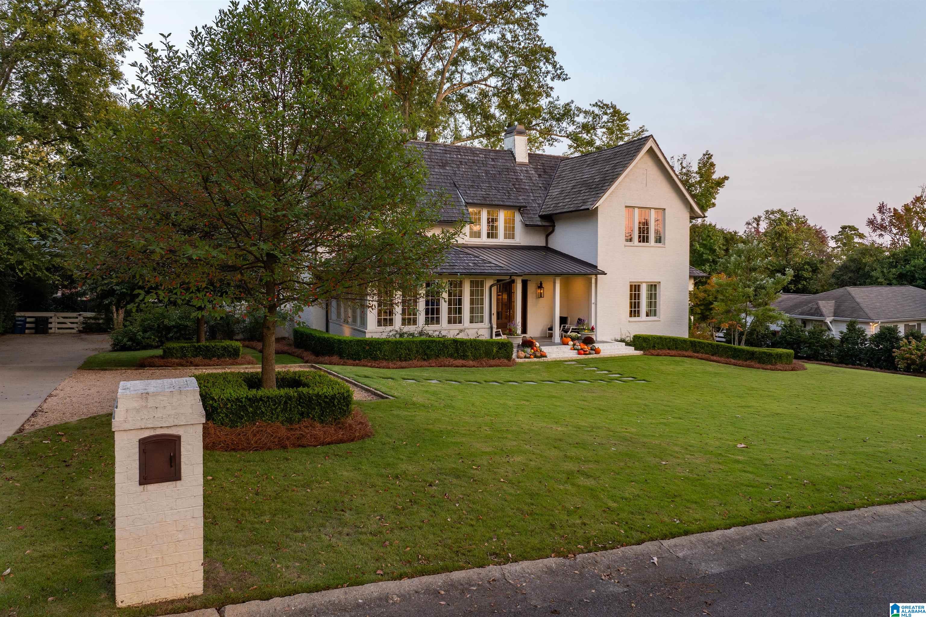 White two-story house with dark roofs, a broad lawn, and pumpkins on the front steps.
