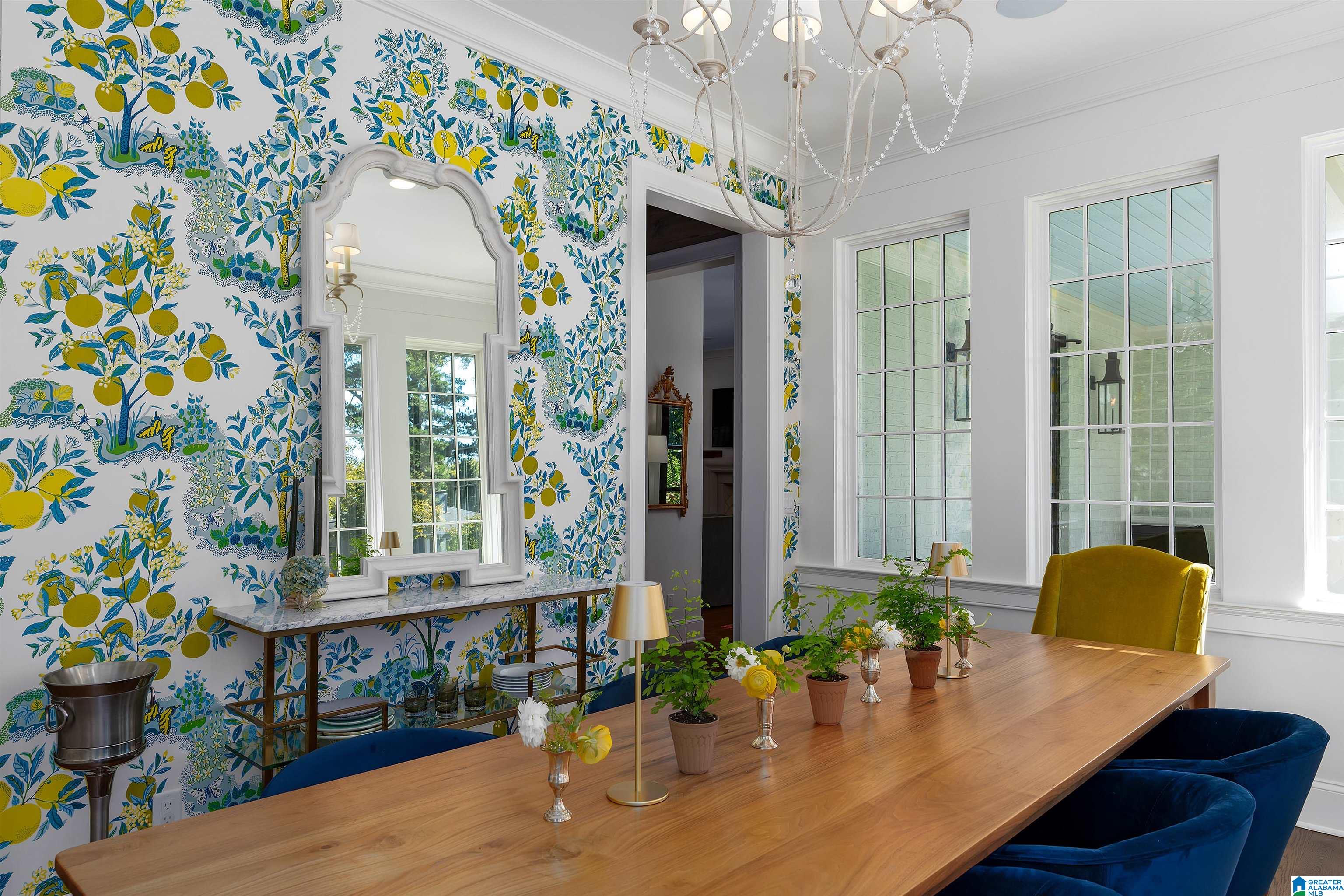 Dining room with citrus-patterned wallpaper, a long wooden table, blue chairs, and tall windows.
