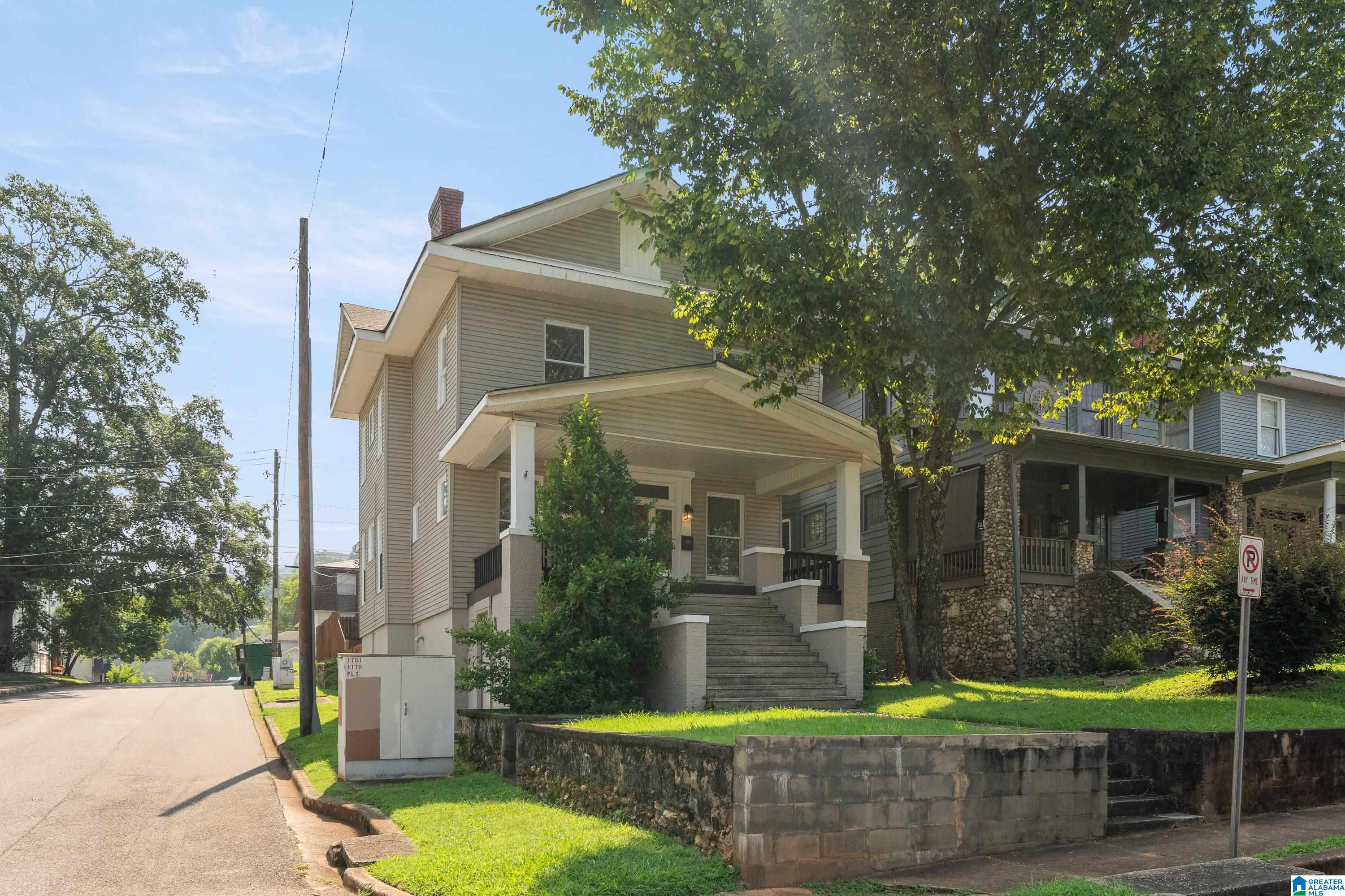 Two-story tan house with a covered porch and front steps, shaded by a large tree.