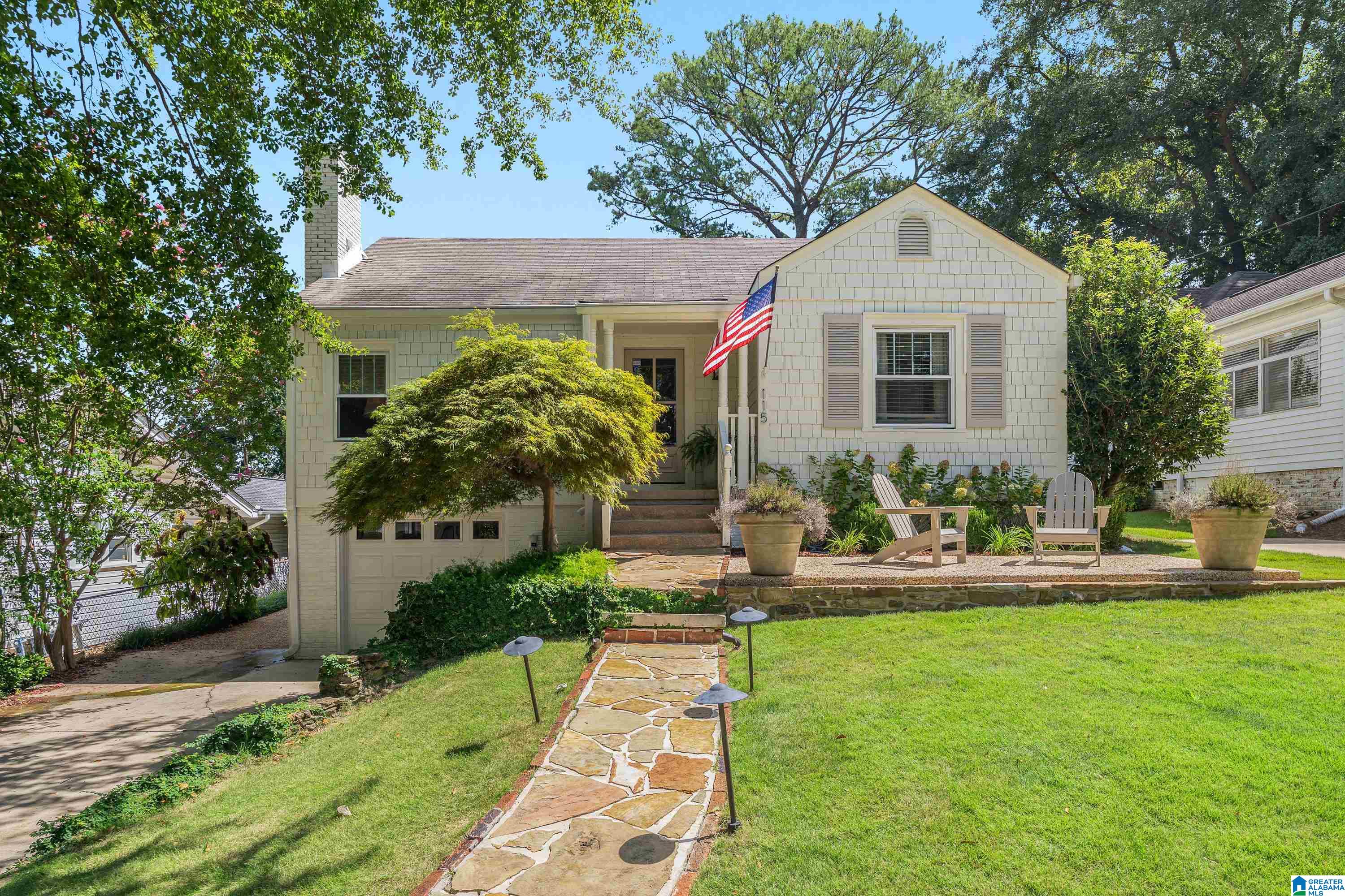 Light shingle house with a U.S. flag, stone walkway, and two Adirondack chairs in the front yard.