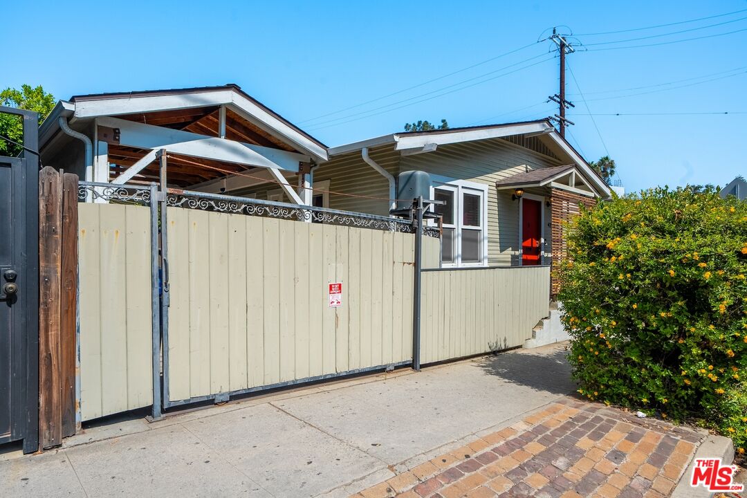Single-story home behind a cream gate, with a red door, covered carport, and flowering hedge.