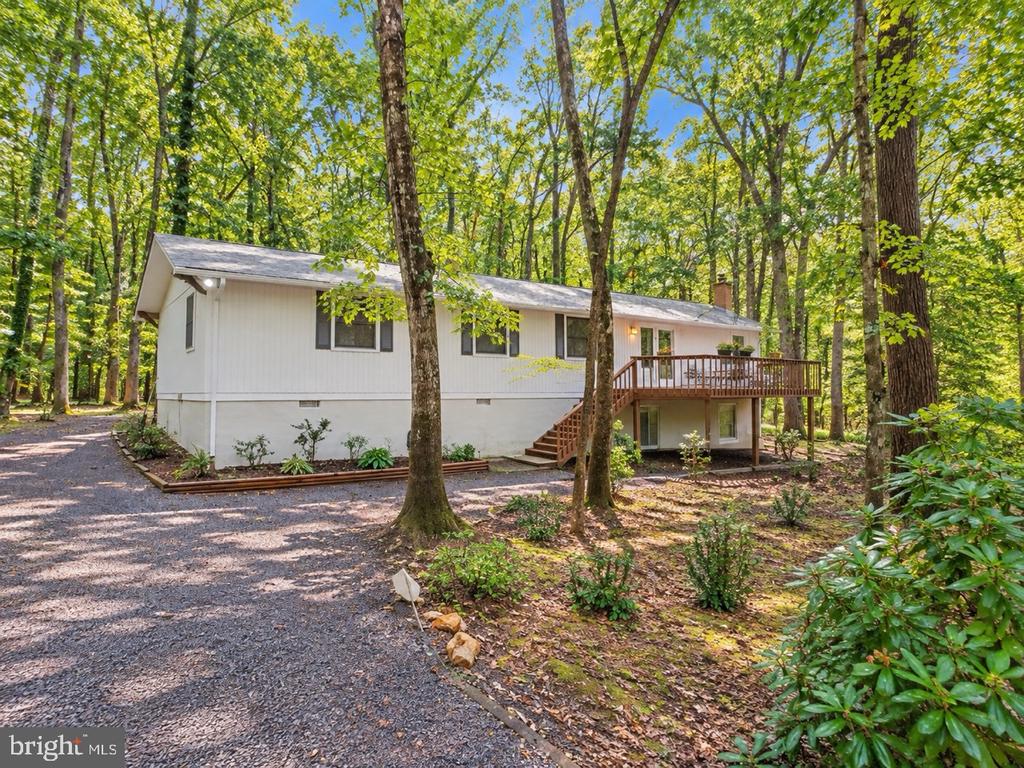 White single-story house in a wooded setting, with a raised deck and gravel driveway.