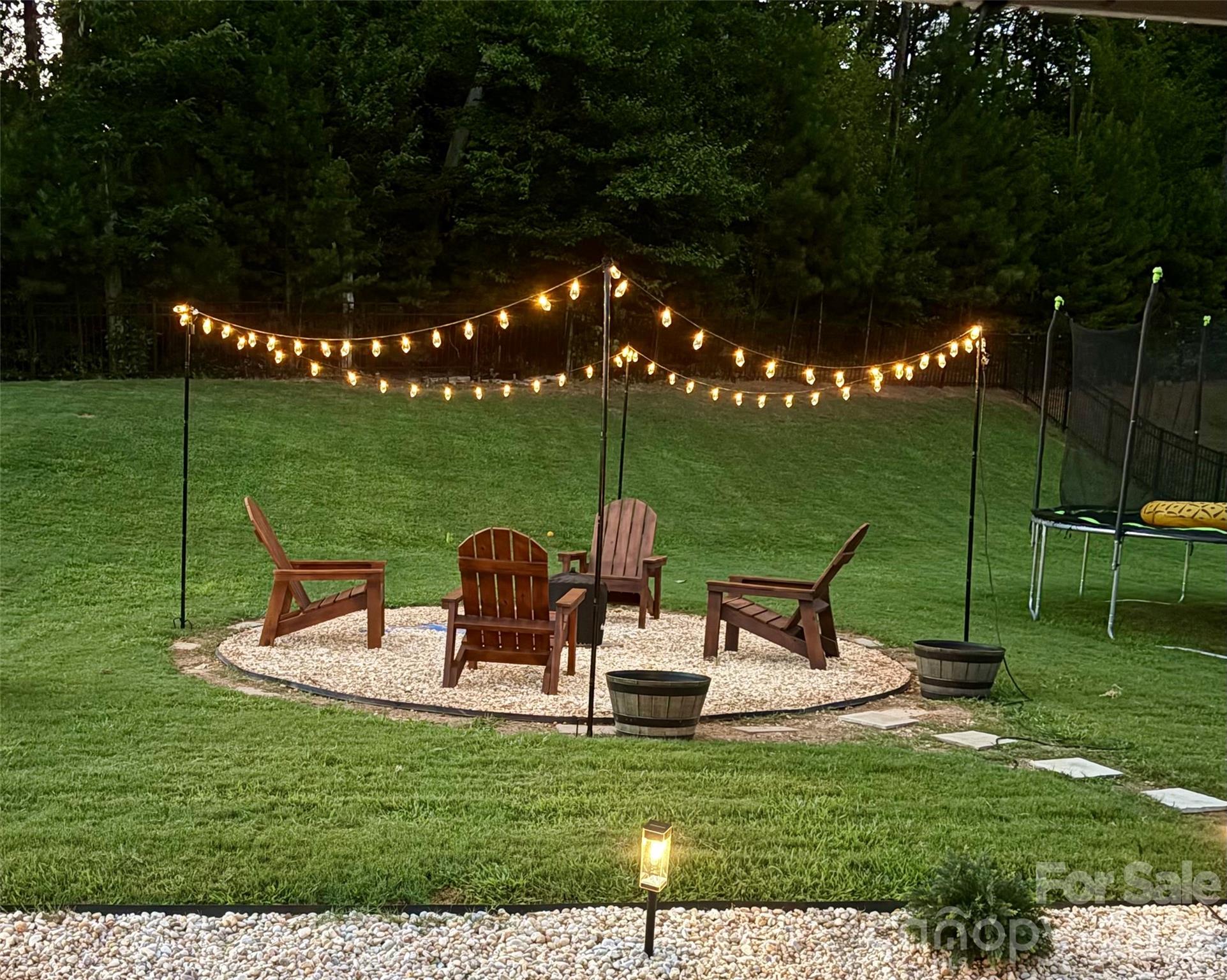 Backyard seating area with Adirondack chairs, glowing string lights, a gravel circle, and a trampoline.