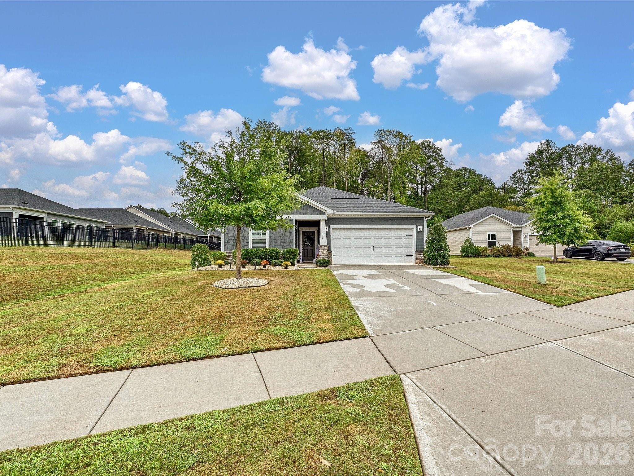 Gray single-story home with a two-car garage, broad driveway, and leafy trees behind it.