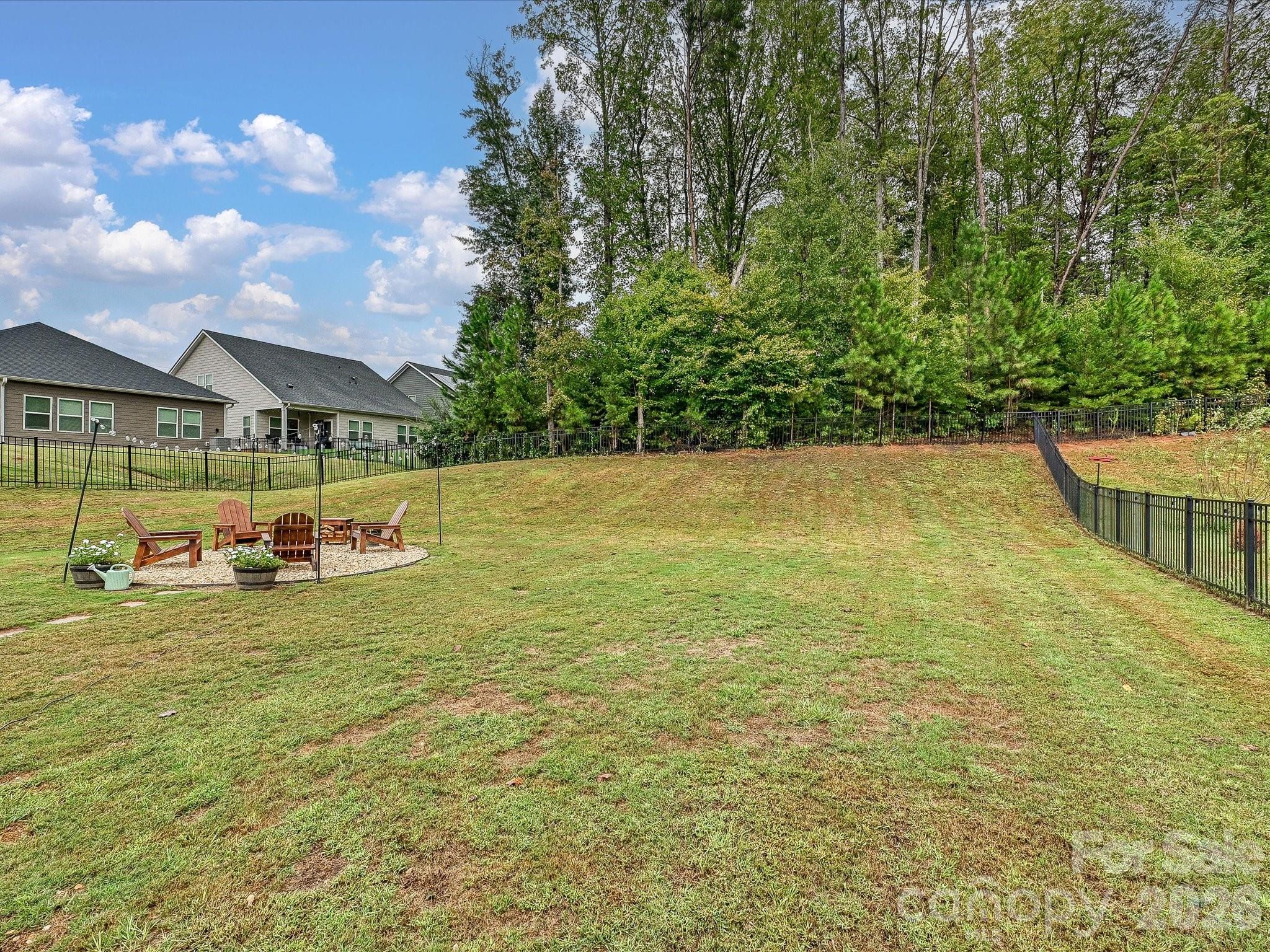 Fenced grassy backyard with a gravel seating area, wooden chairs, and mature trees along the rear boundary.