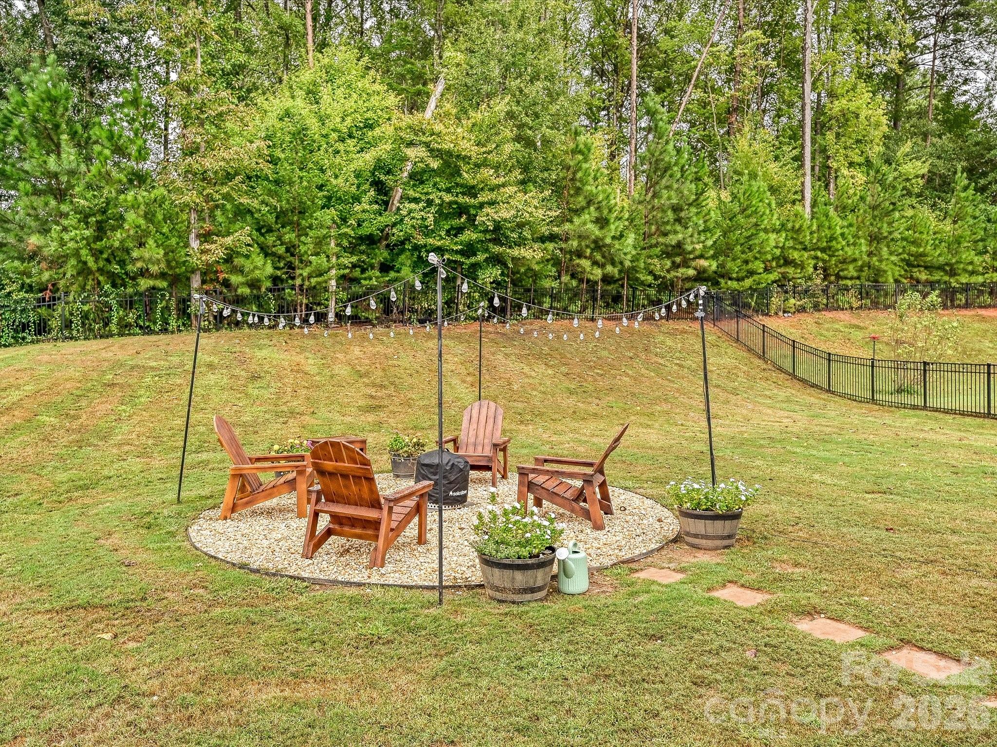 Backyard fire pit seating area with wooden Adirondack chairs, overhead string lights, and a wooded backdrop.
