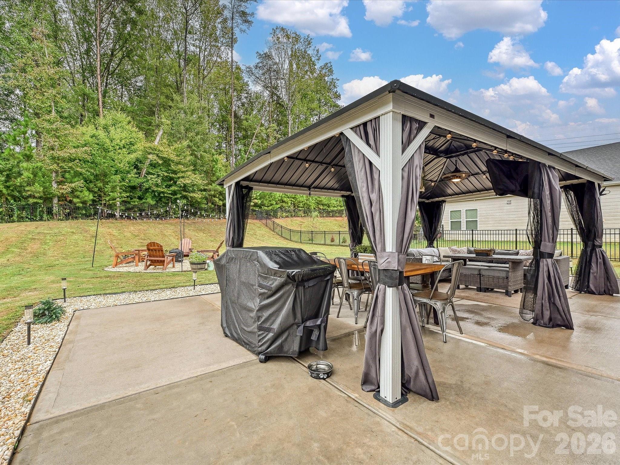 Covered backyard patio with curtained gazebo, dining and lounge seating, and a wooded lawn.