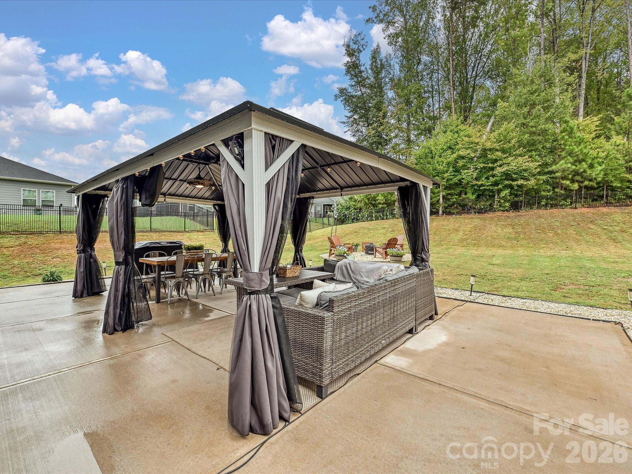 Covered backyard gazebo with tied-back curtains, a dining set, and wicker lounge seating on a concrete patio.
