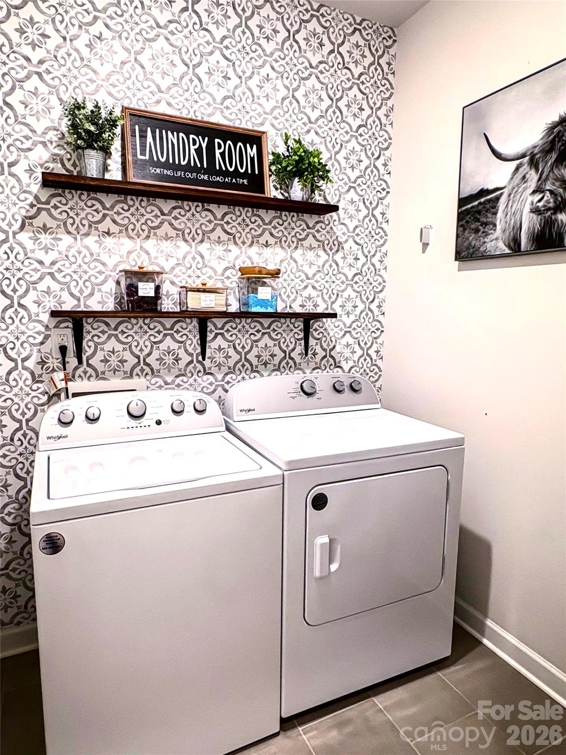 Laundry room with a white washer and dryer, patterned wallpaper, and dark wood display shelves.