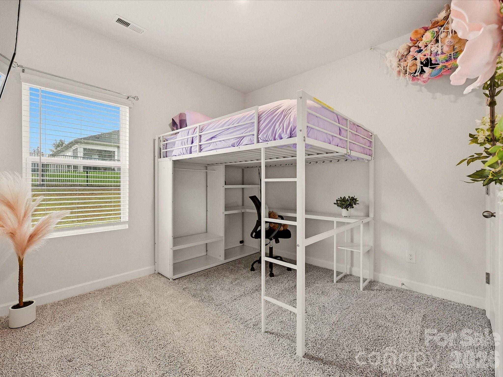 Bedroom with a white loft bed, lavender bedding, and a desk underneath beside a window.