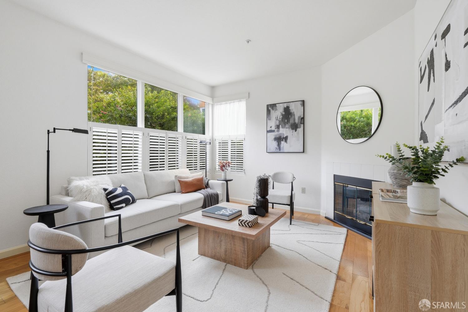 Bright living room with a white sofa, corner fireplace, and large windows overlooking greenery.