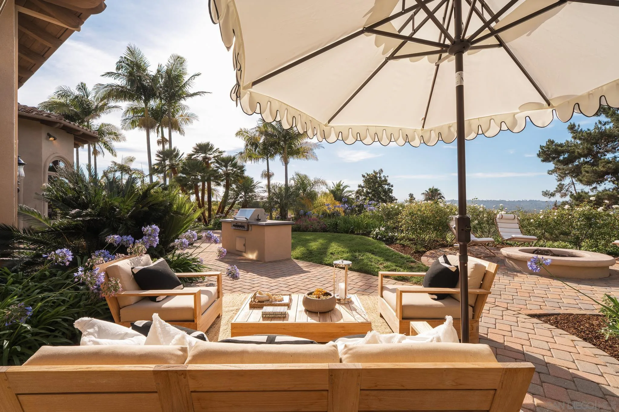Outdoor patio with cushioned seating, a large umbrella, built-in grill, and circular fire pit amid tropical landscaping.