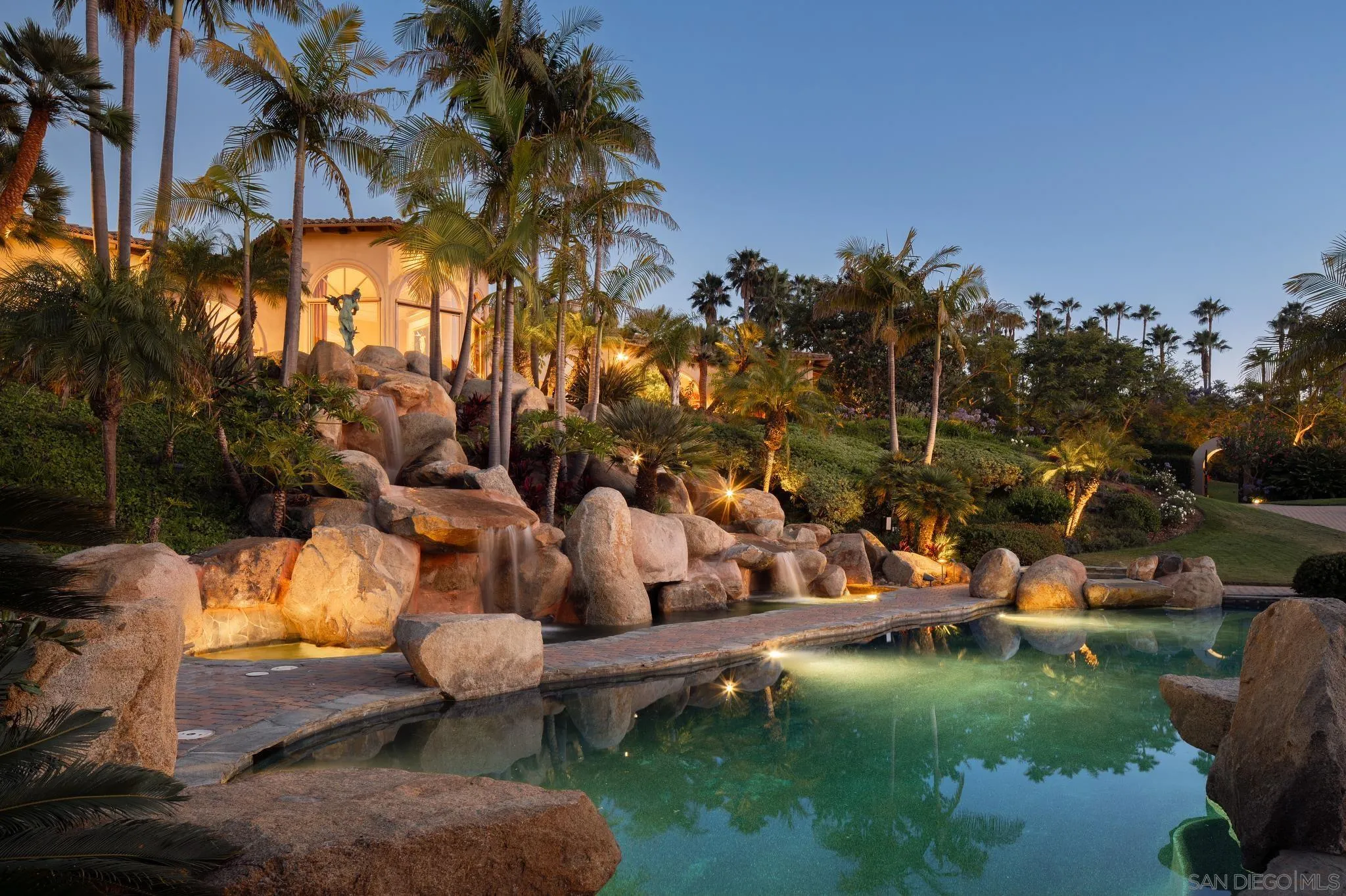 Estate pool with boulder waterfalls, palm trees, and warmly lit house at dusk.