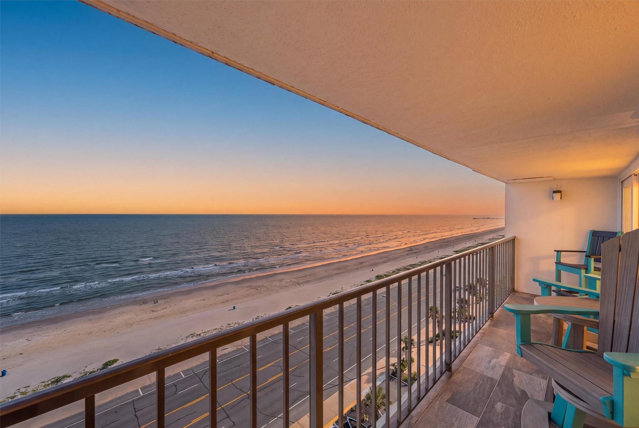 Oceanfront balcony at sunset overlooking a sandy beach, with turquoise chairs beside the railing.