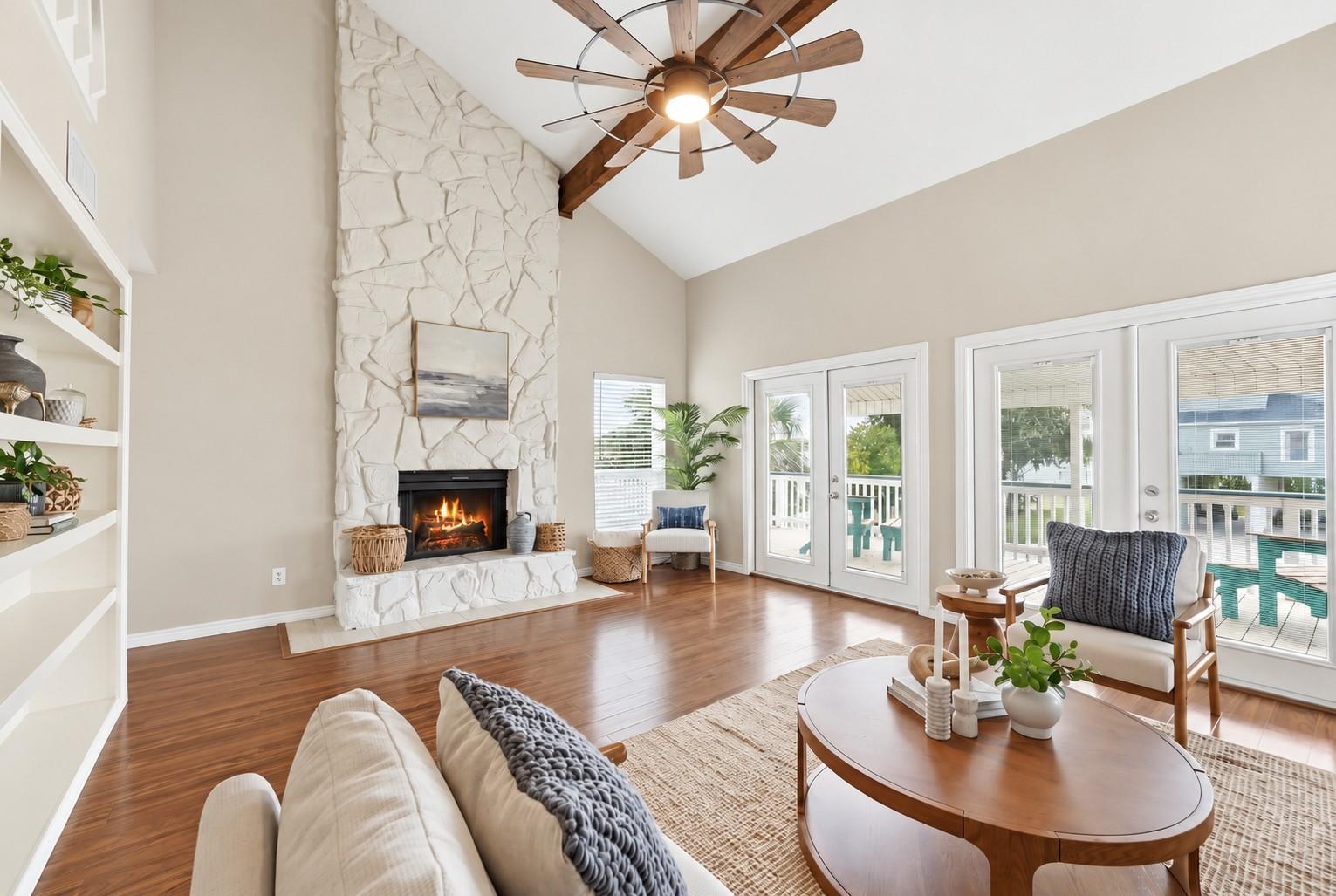 Living room with a vaulted ceiling, white stone fireplace, hardwood floors, and doors to a deck.