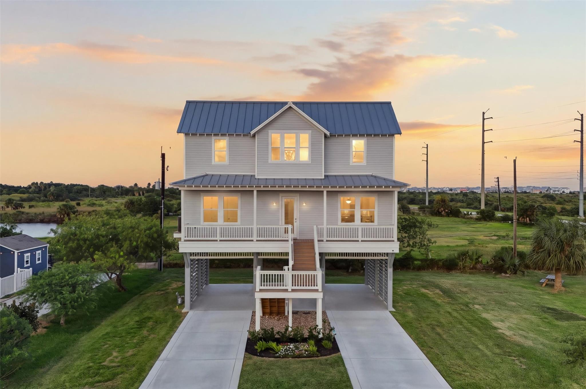 Raised gray two-story house with a blue metal roof and broad front porch at sunset.