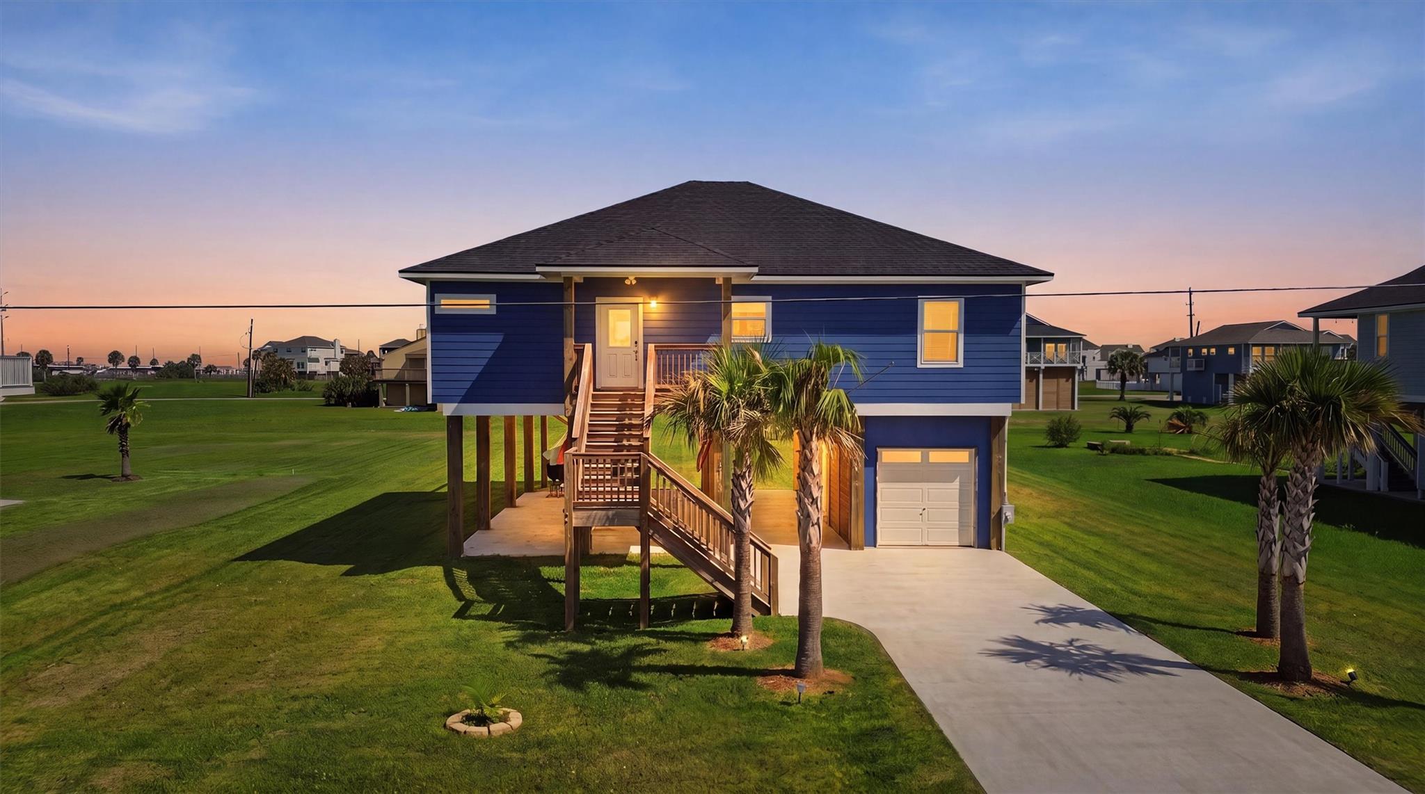 Blue elevated house with a raised porch, palm trees, and glowing windows at dusk.