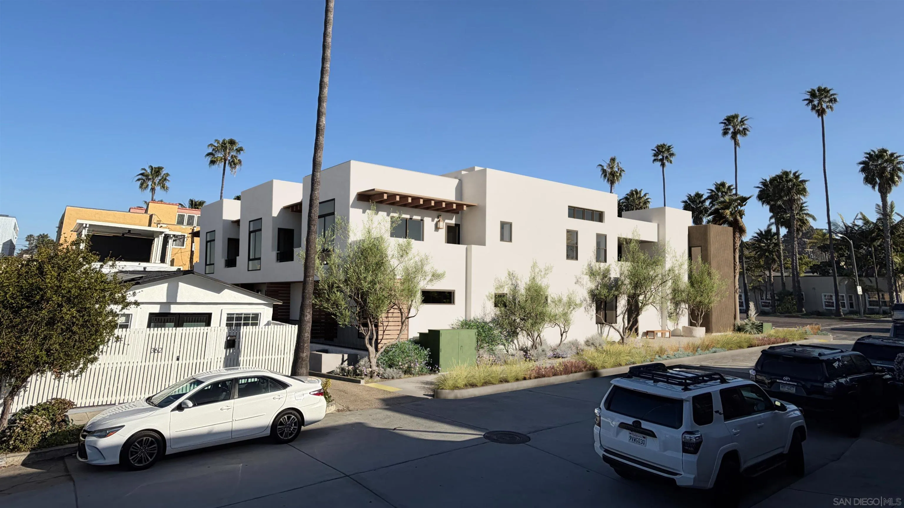 Modern white residential building with angular architecture, landscaped frontage, and tall palm trees.