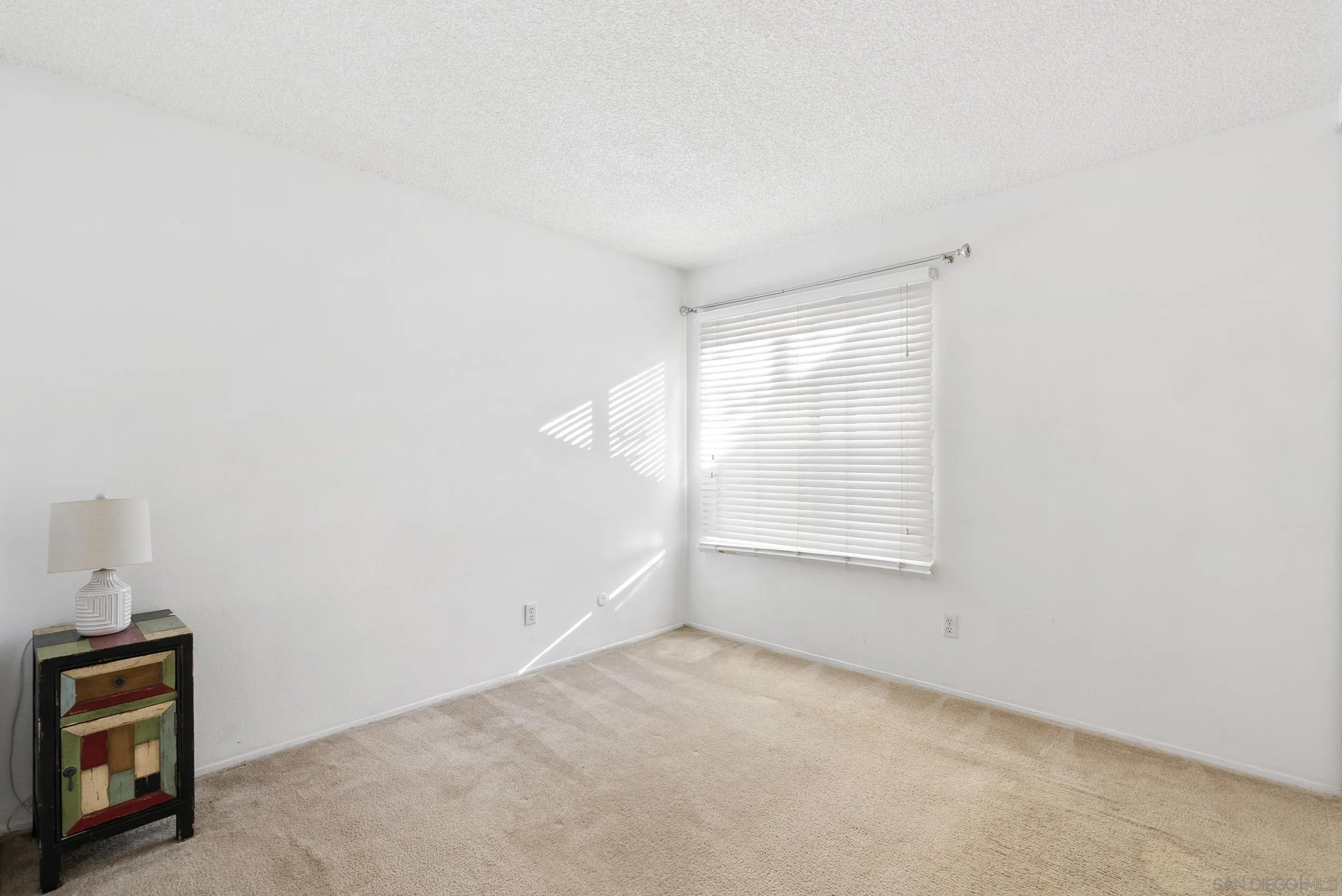 Sparse bedroom with beige carpet, white walls, closed window blinds, and a colorful cabinet with a lamp.