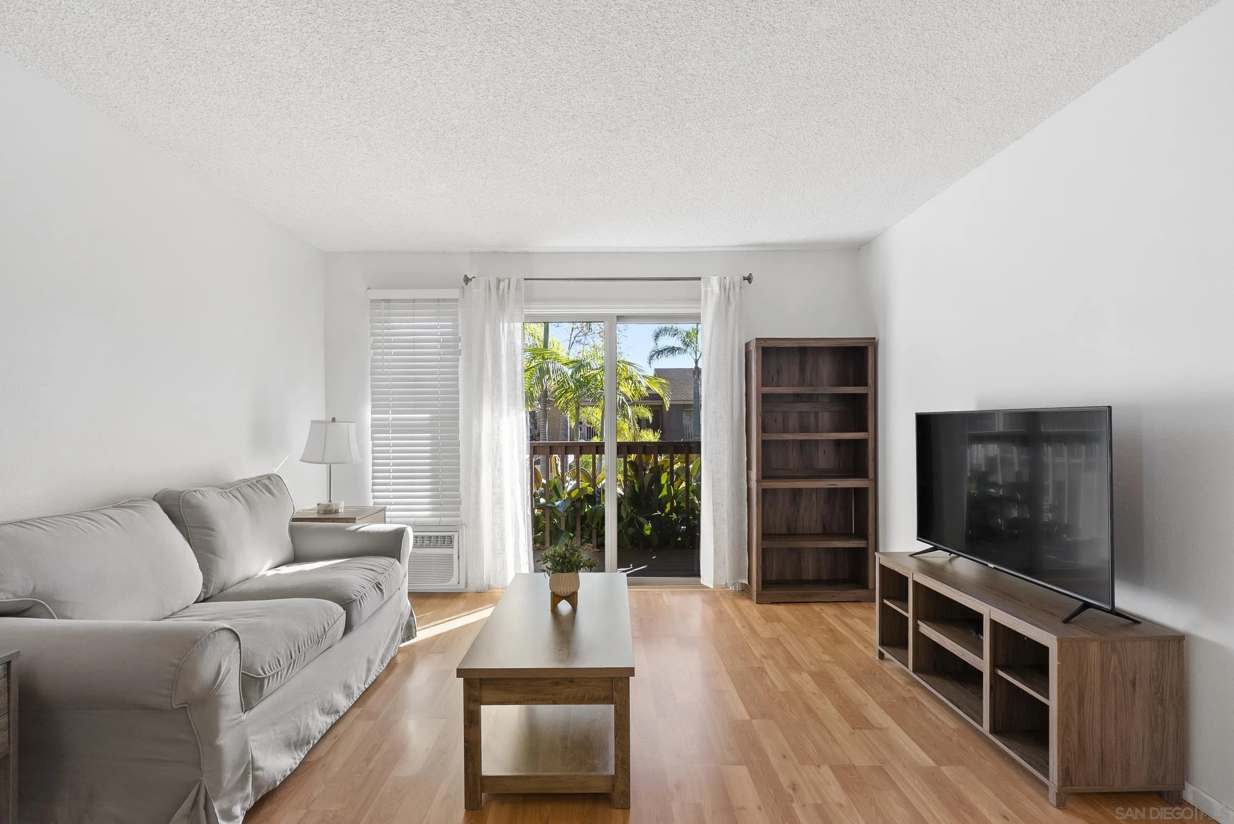 Living room with a gray sofa, wood furnishings, and sliding doors overlooking leafy patio plants.