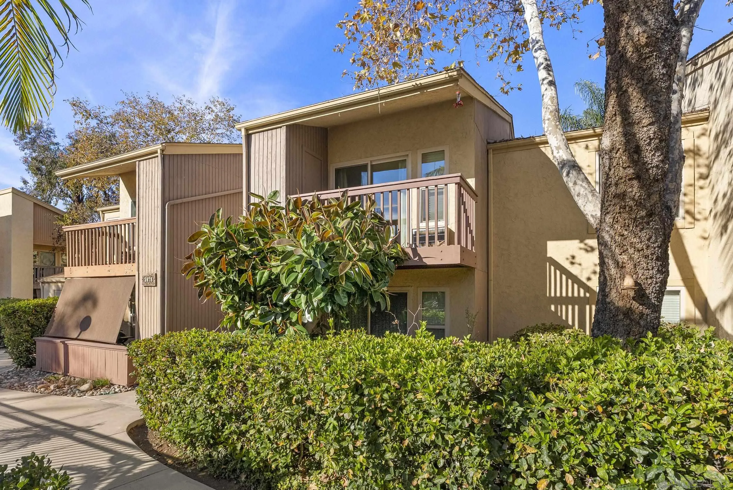 Two-story condominium exterior with brown balconies, dense hedges, and a large shade tree.