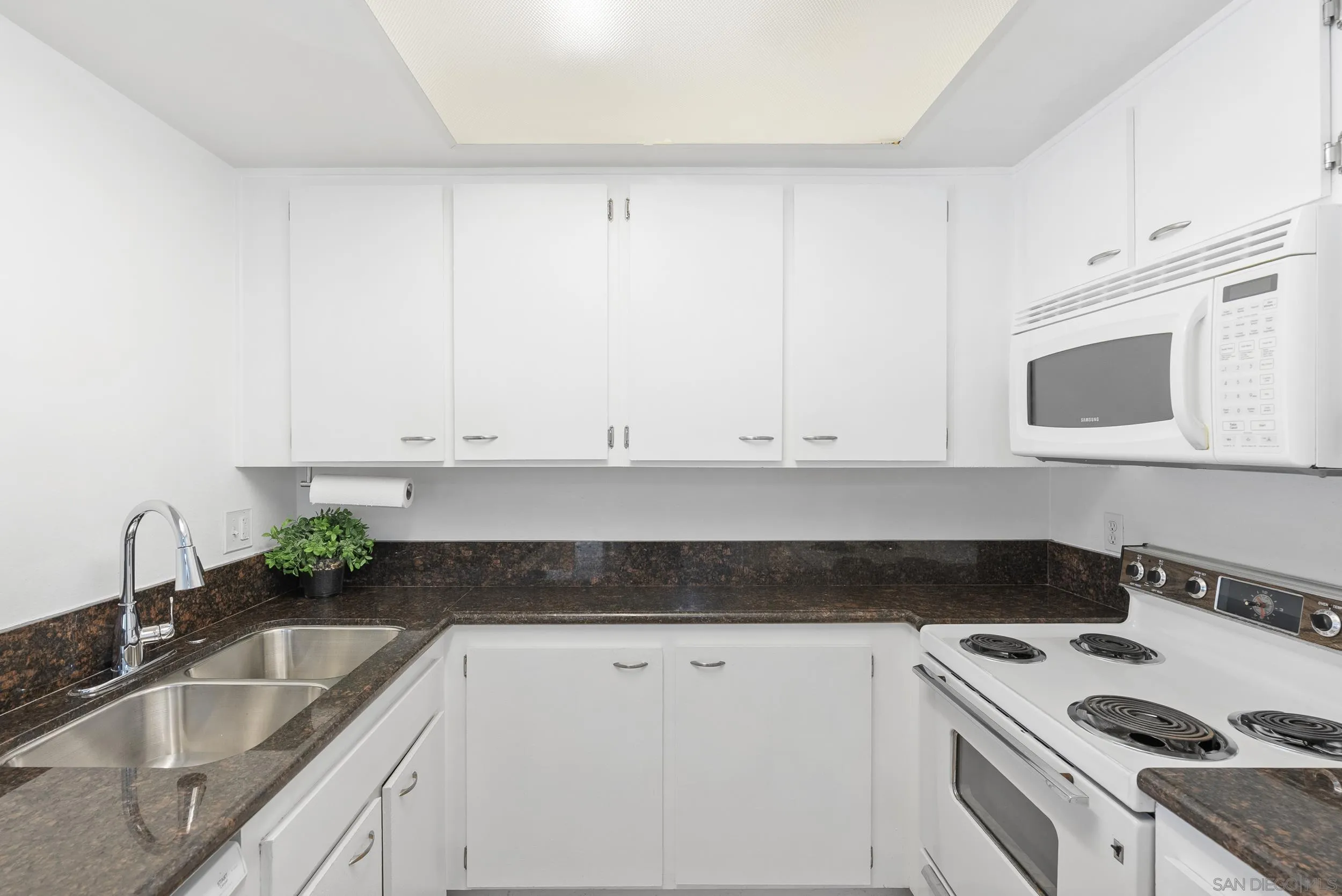 Compact kitchen with white cabinets, dark stone counters, a double sink, and white electric appliances.