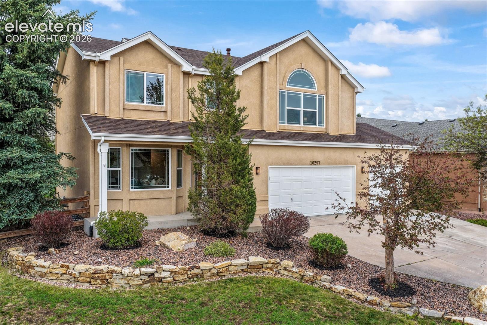 Two-story tan stucco home with an arched upper window, attached garage, and stone-edged landscaping.