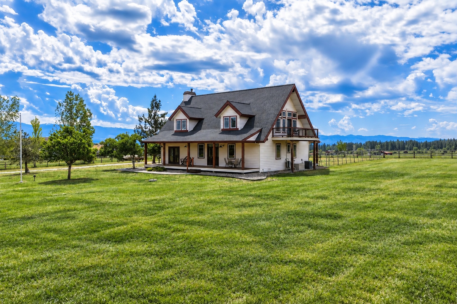Two-story white house with a covered porch and balcony on a broad lawn, with mountains in the distance.