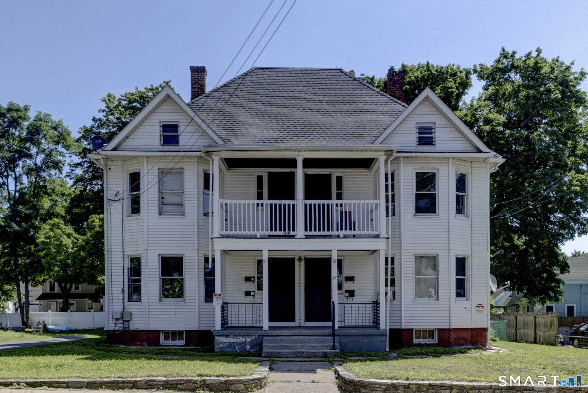 White two-story duplex with two porches, separate entrances, and a stone-edged front lawn.