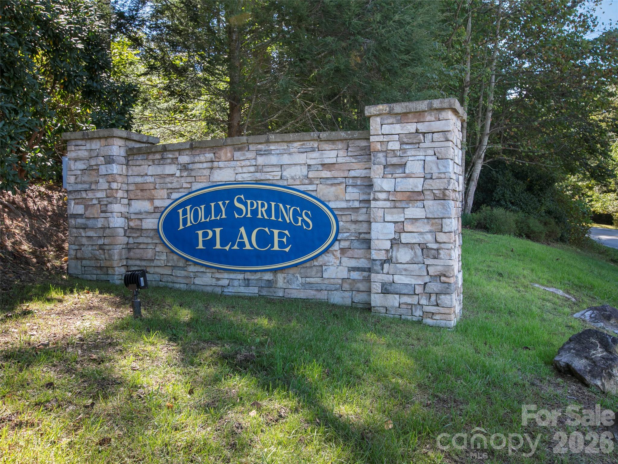 Stone entrance sign for Holly Springs Place beside a grassy lawn and wooded landscaping.
