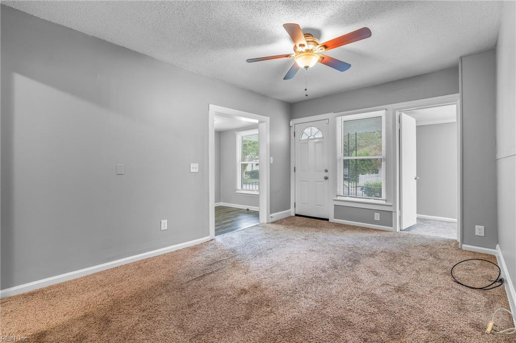 Empty living room with beige carpet, gray walls, and a lit ceiling fan near the front door.