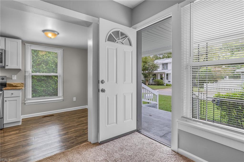 Front entry with open door to a covered porch, carpeted floor, and large windows with blinds.