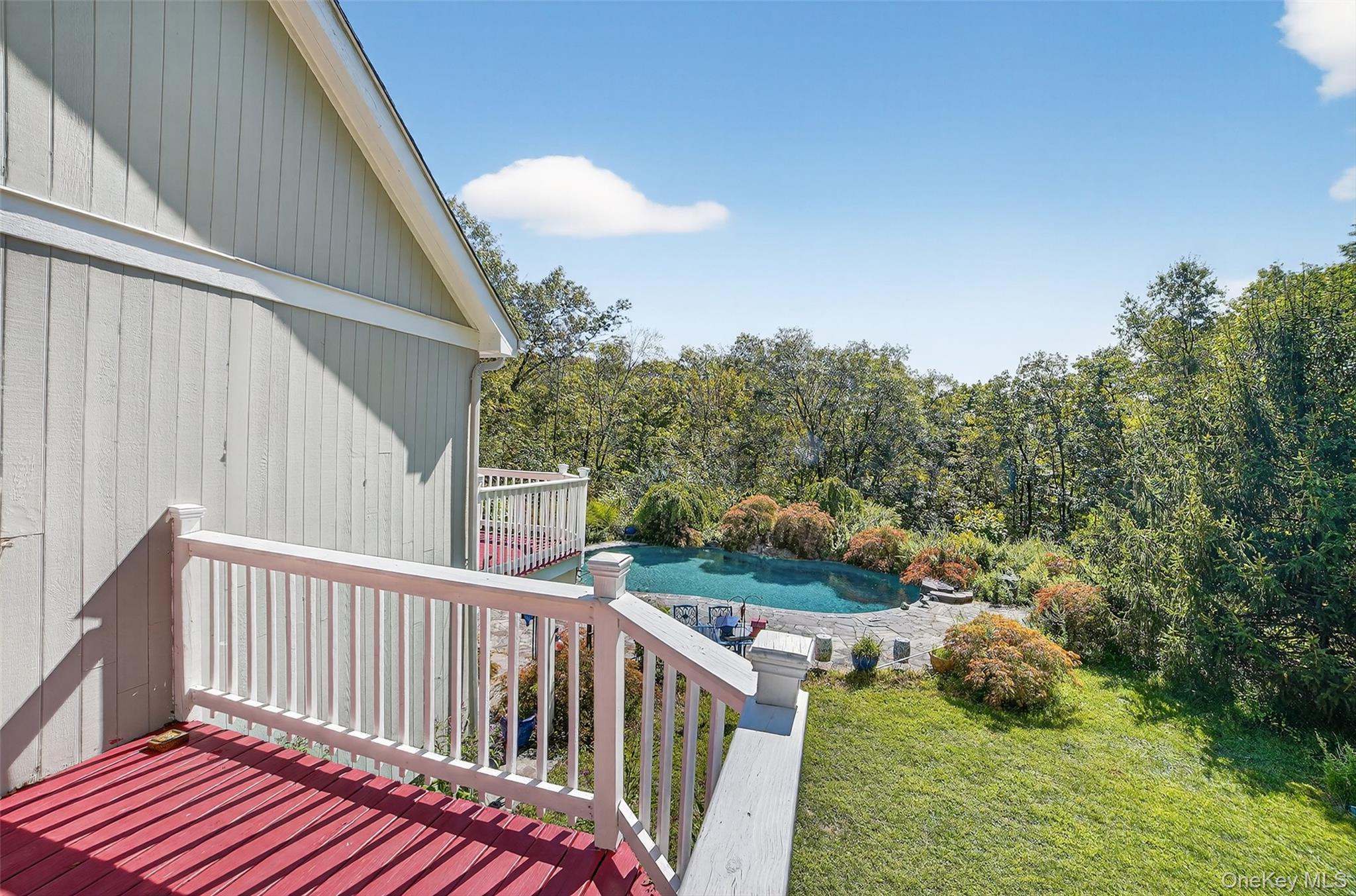 Red deck overlooks a swimming pool and stone patio surrounded by trees.