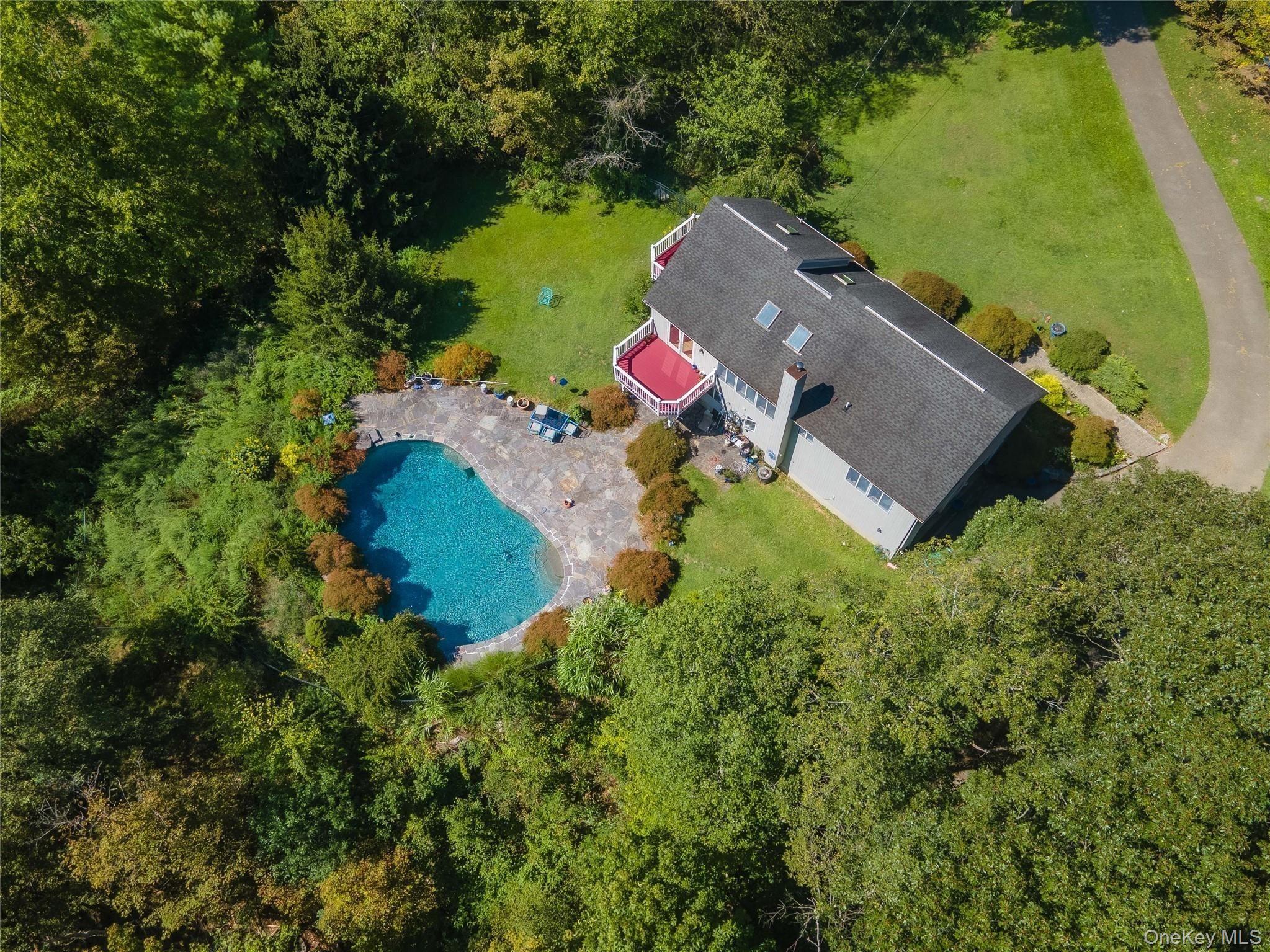 Aerial view of a house with a stone patio, swimming pool, red deck, and wooded grounds.