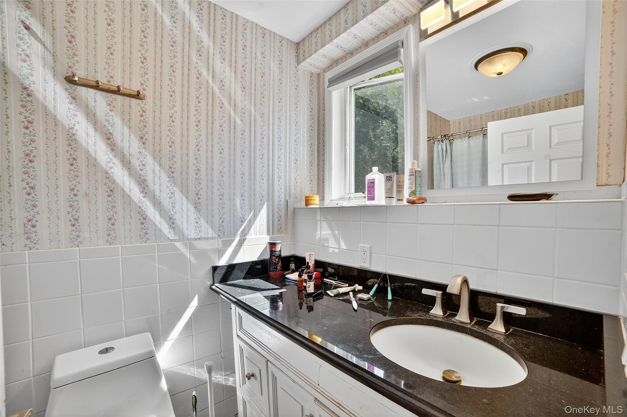 Bathroom with a black countertop, white vanity and toilet, floral wallpaper, and sunlight through the window.