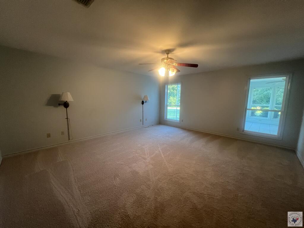 Empty carpeted living room with a ceiling fan, windows, and glass door to a tiled sunroom.