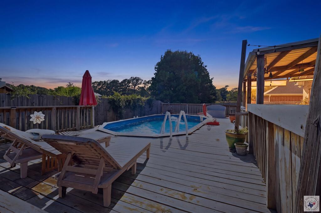 Backyard pool with wood deck, lounge chairs, and a covered patio at dusk.